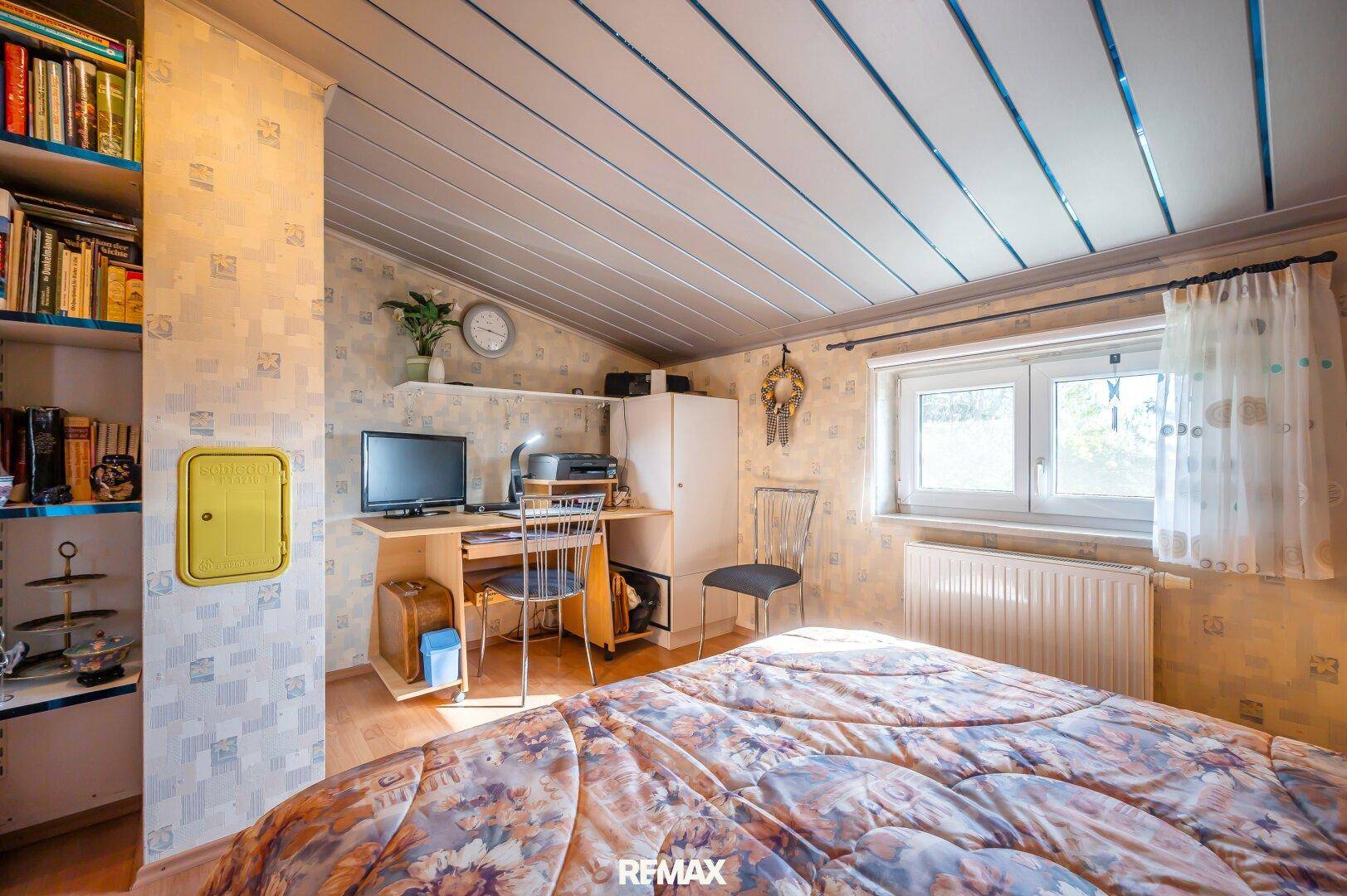 Schlafzimmer mit Dachschräge, Holzboden und Arbeitsbereich am Fenster.