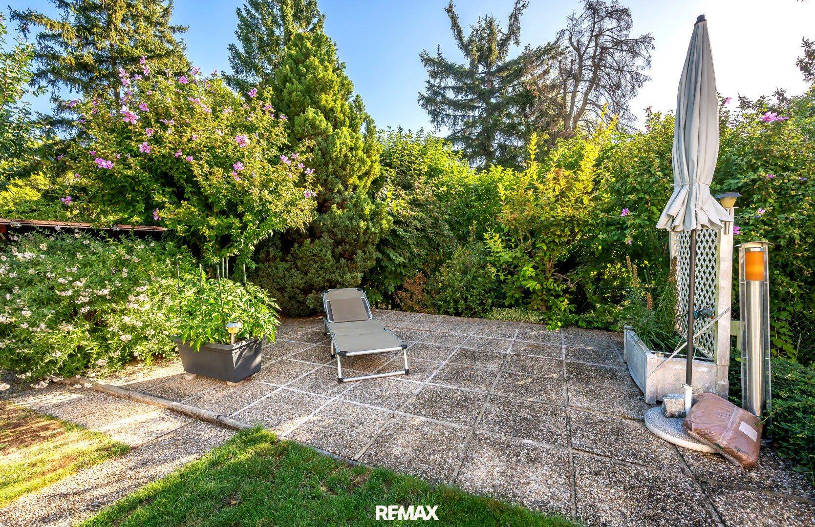 Sonniger Terrassenbereich im Garten mit Liegestuhl und üppiger Bepflanzung.