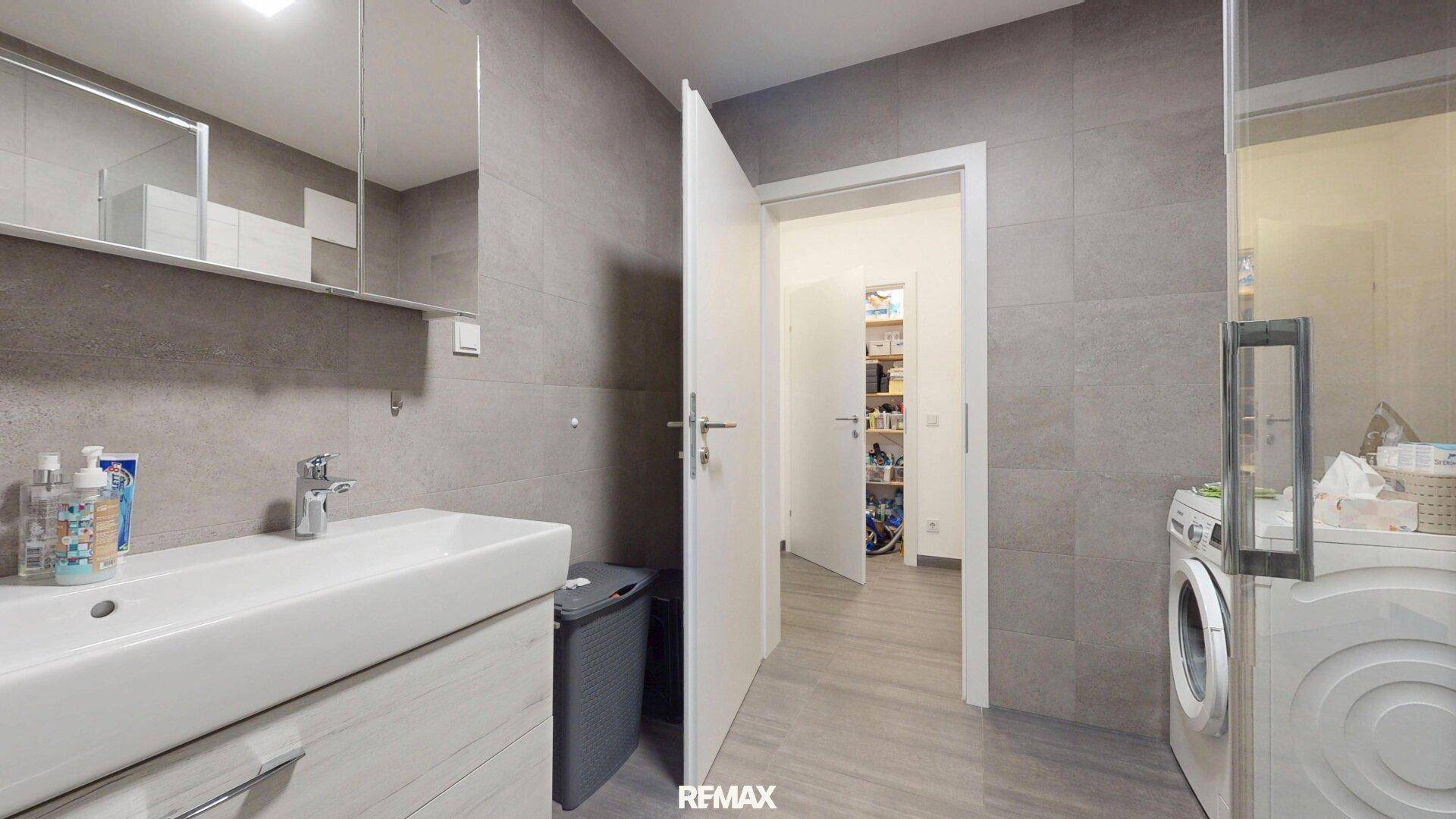 Modernes Badezimmer mit grauen Fliesen, Waschbecken und Blick in einen praktischen Abstellraum.