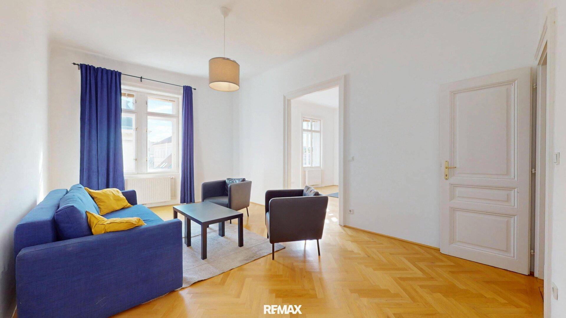 Helles Wohnzimmer mit blauem Sofa, Parkettboden und Zugang zu weiteren Räumen.