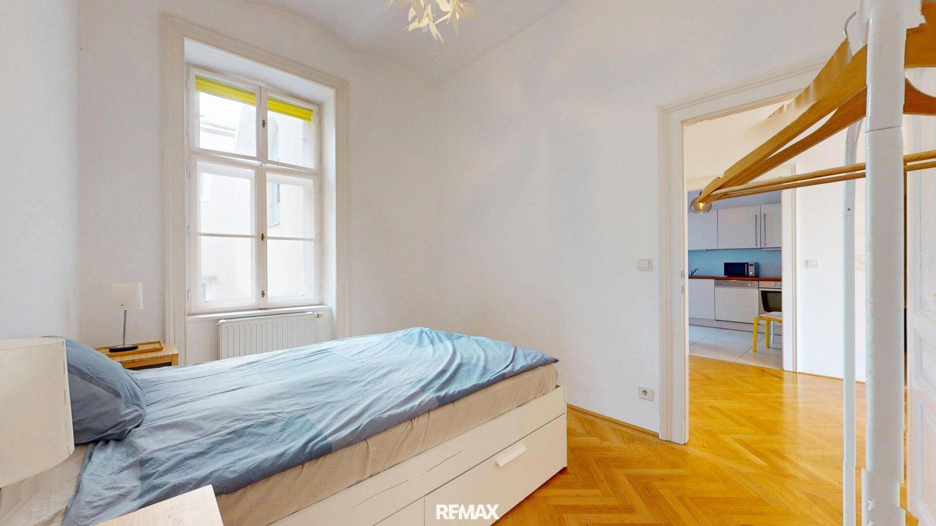 Helles Schlafzimmer mit Parkettboden und Blick in die angrenzende Küche.