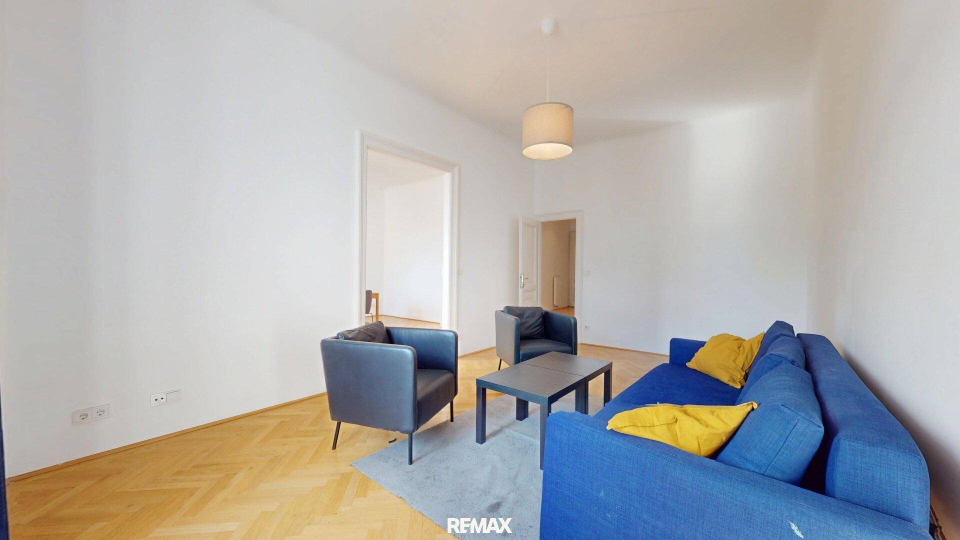 Modern eingerichtetes Wohnzimmer mit blauer Couch und dunklen Sesseln auf hellem Parkettboden.
