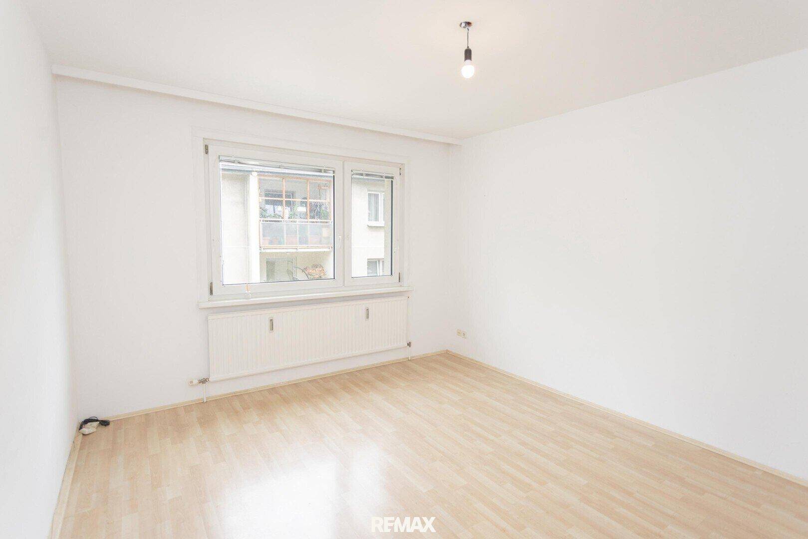 Geräumiges, helles Zimmer mit Laminatboden und Blick nach draußen durch das Fenster.