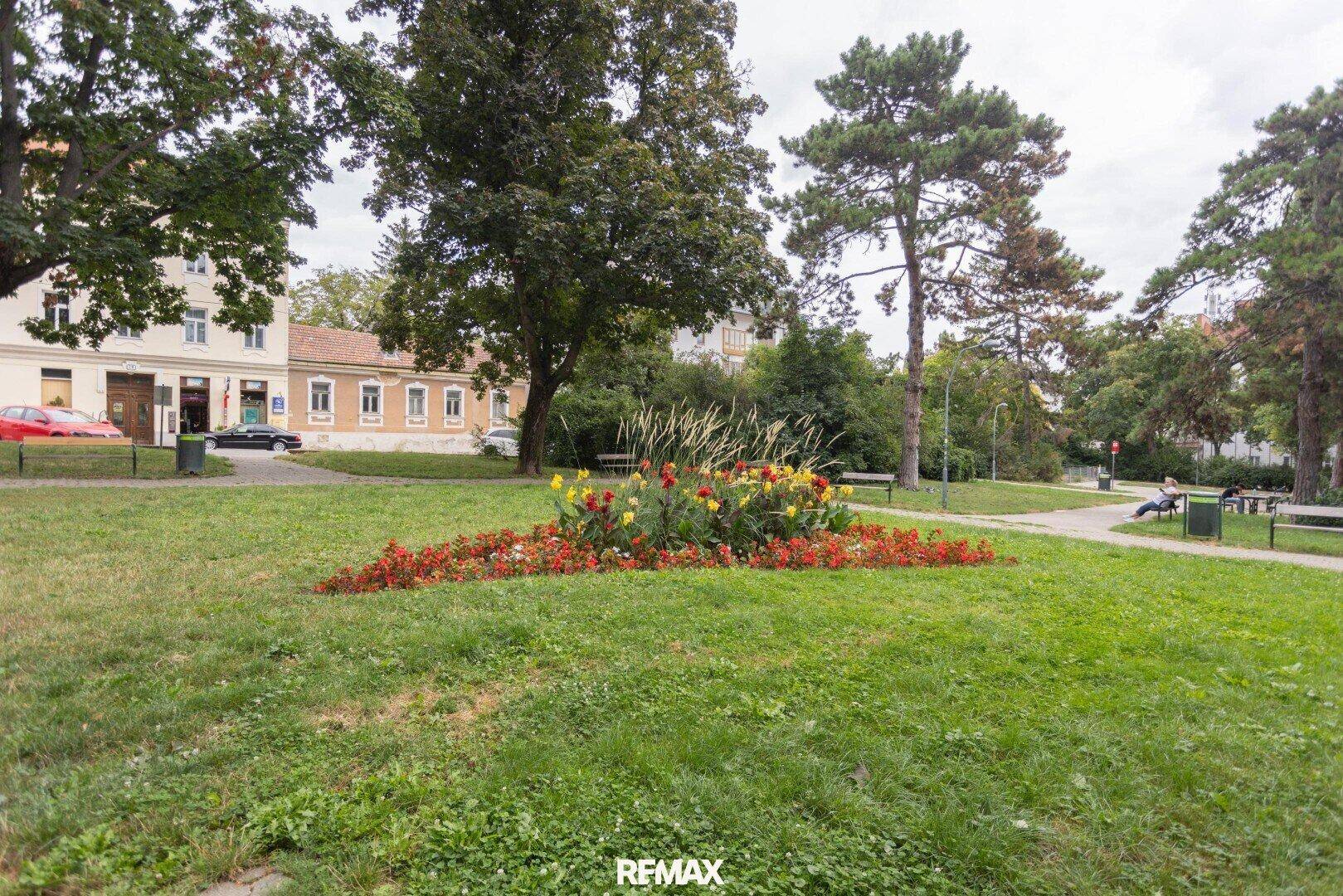Grüner Park mit Blumenbeet und Bäumen, umgeben von Wohngebäuden.