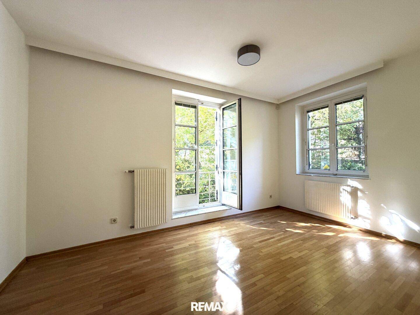 Heller Raum mit Parkettboden und zwei Fenstern, eines davon als Fenstertür mit Blick ins Grüne.