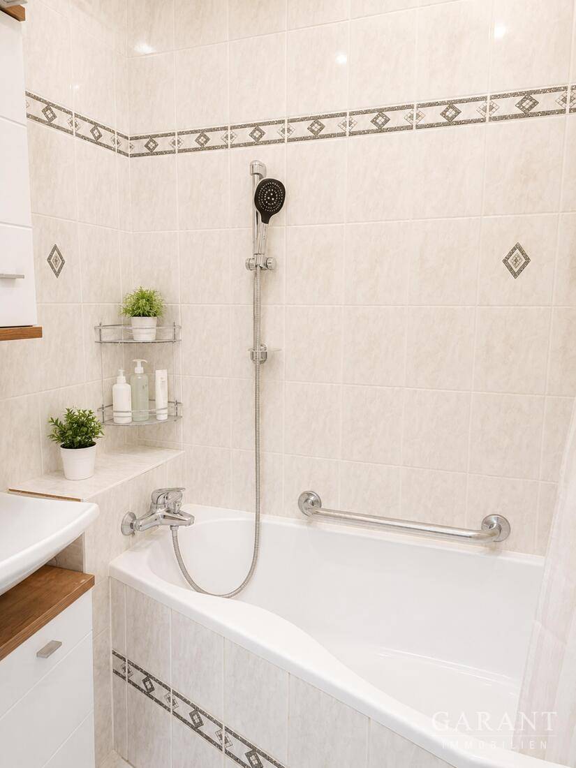 Badezimmer mit Badewanne, Duscharmatur und hellen Fliesen mit dekorativen Akzenten.