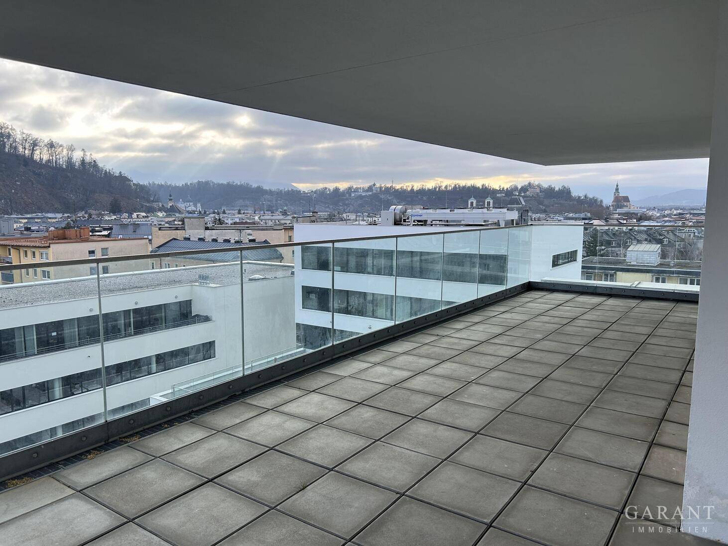 Große Dachterrasse mit Fliesenboden und Glasgeländer bietet eine weite Aussicht über die Stadt.