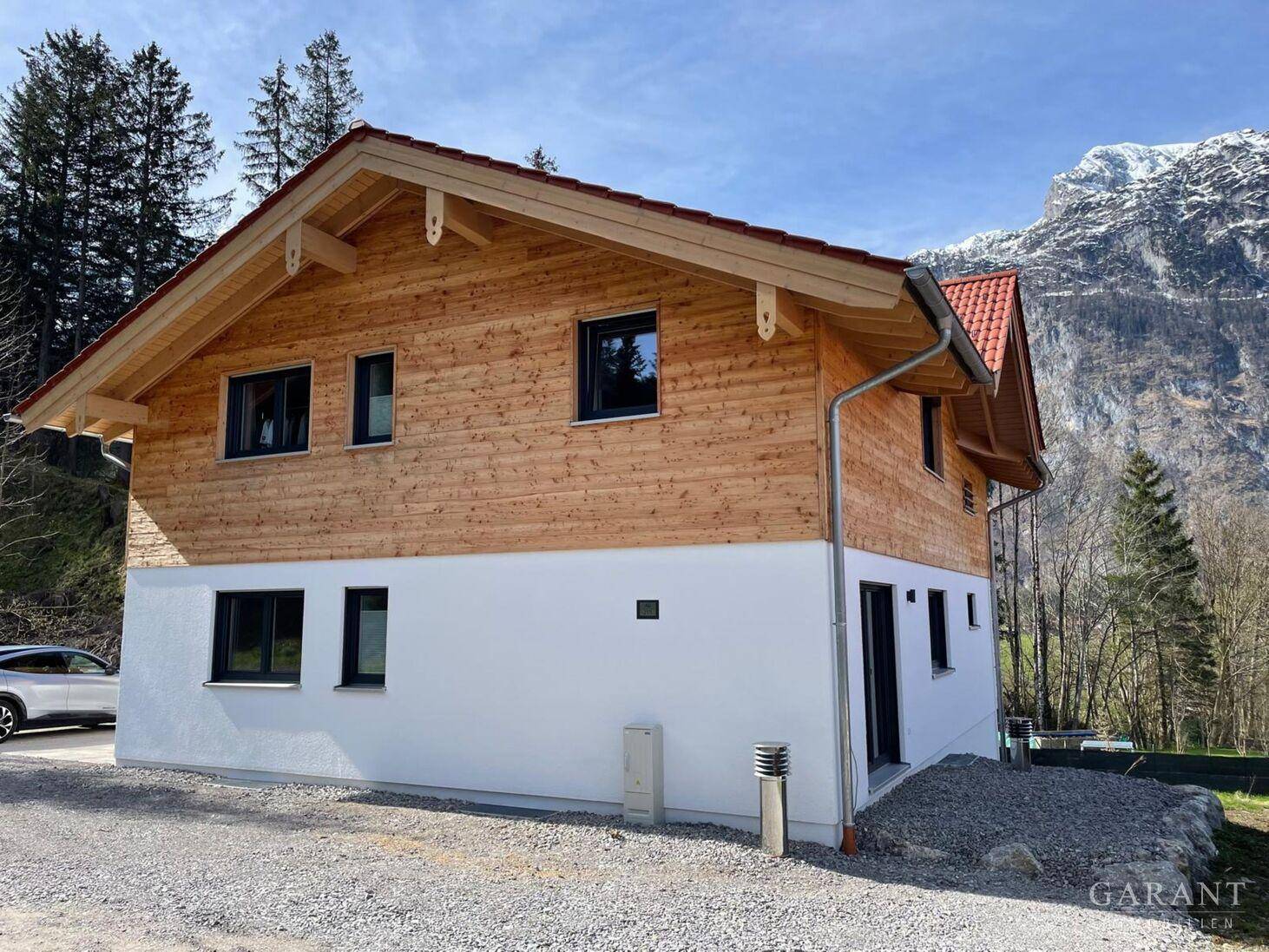 Neubau-Holzhaus mit weißem Sockel und rotem Dach vor beeindruckender Bergkulisse.