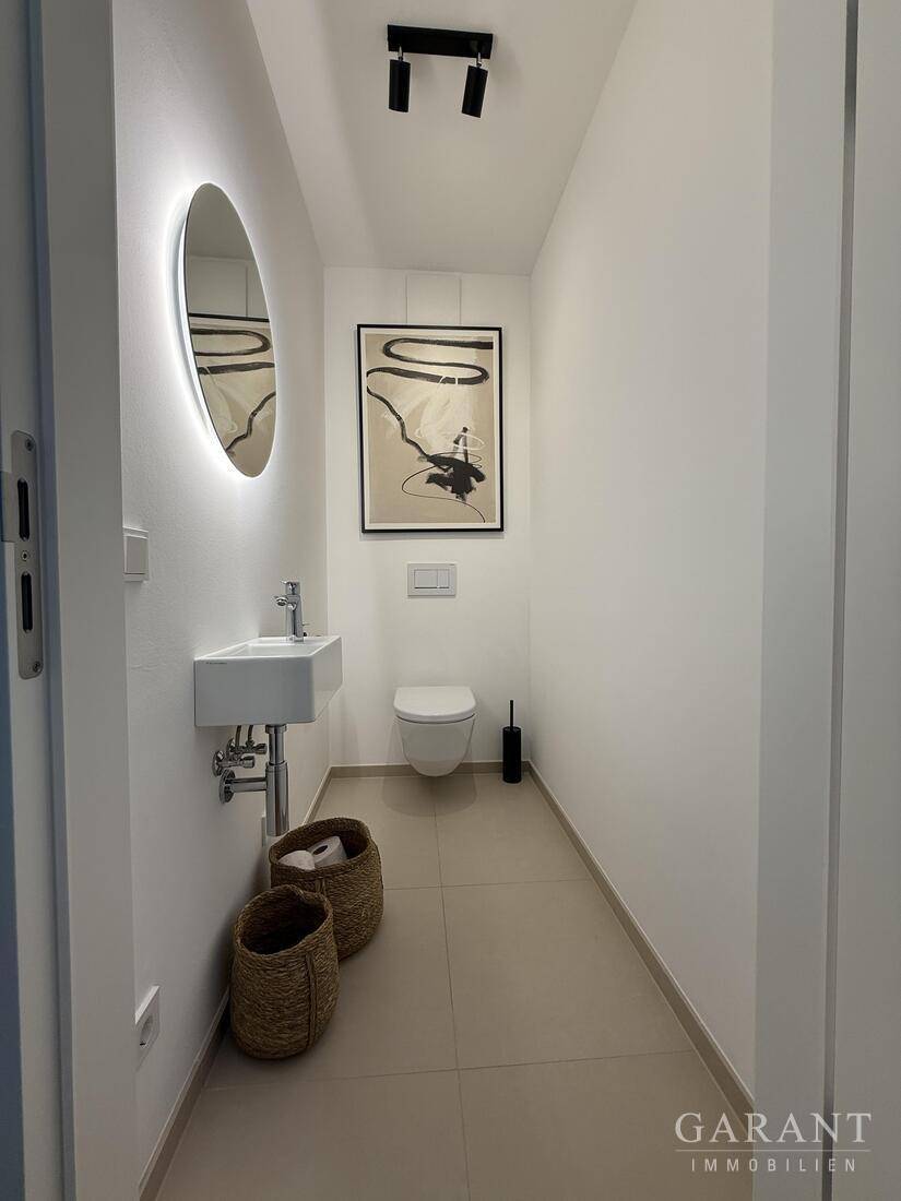 Modernes Gäste-WC mit wandmontierter Toilette, kleinem Waschbecken und beleuchtetem Spiegel.