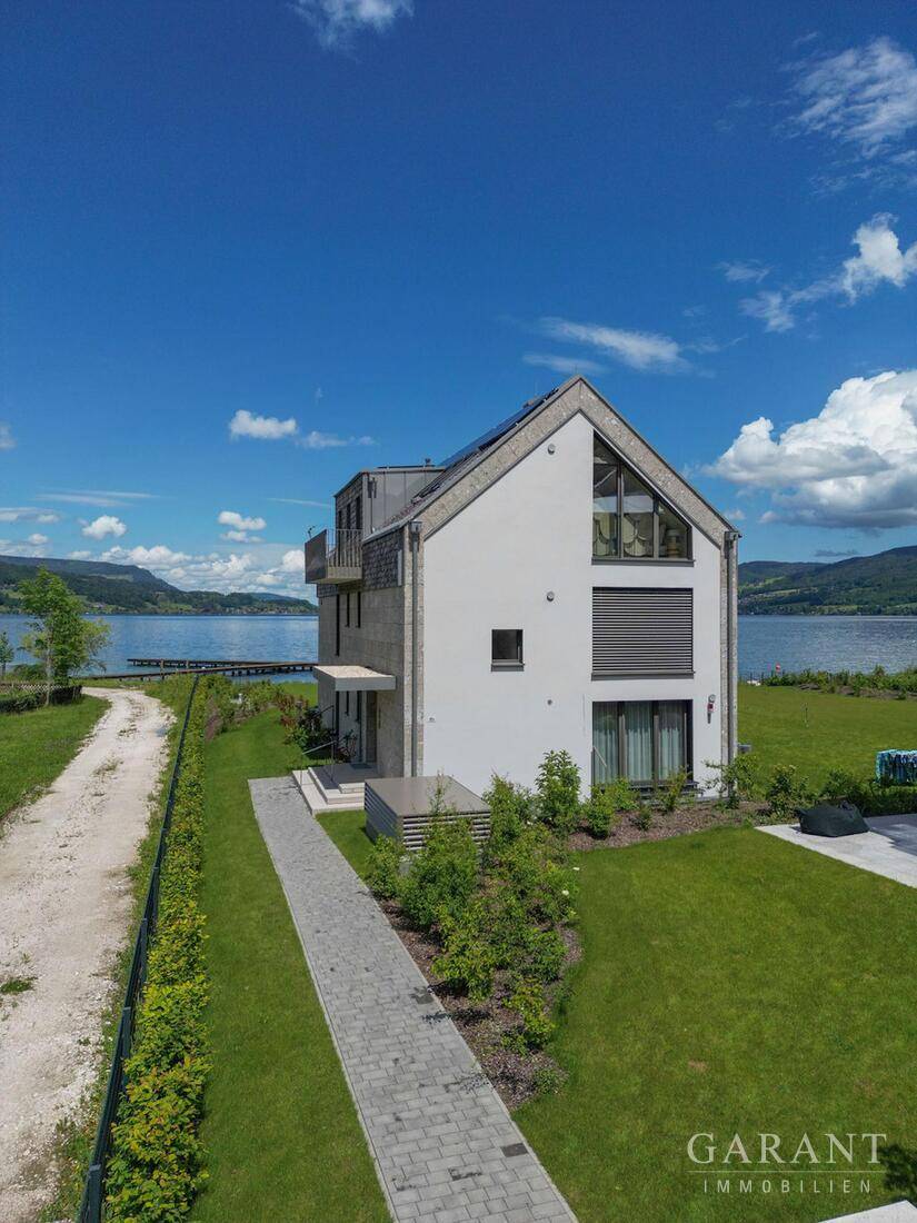 Modernes Gebäude mit gepflegtem Garten und direktem Zugang zum See unter blauem Himmel, sehr idyllisch.