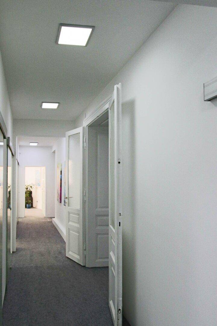 Heller Bürogang mit grauen Teppichboden, weißen Türen und quadratischen Deckenleuchten.