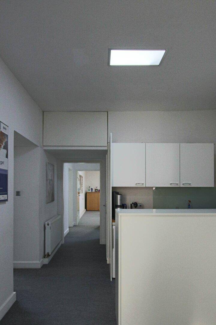 Heller Flur mit integrierter Küchenzeile, grauen Teppichboden und Deckenleuchten.