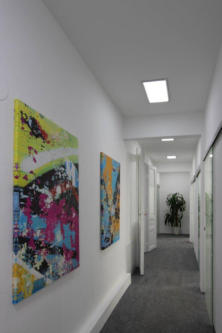 Langer, heller Flur mit grauen Teppichboden, modernen Kunstwerken und mehreren Türen zu Büroräumen.