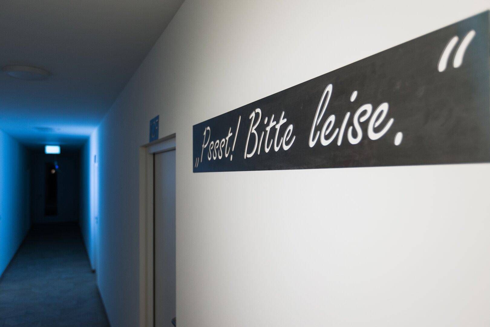 Langer, schmaler Flur mit weiß gestrichenen Wänden und einem Schild mit der Aufschrift "Psst! Bitte leise."