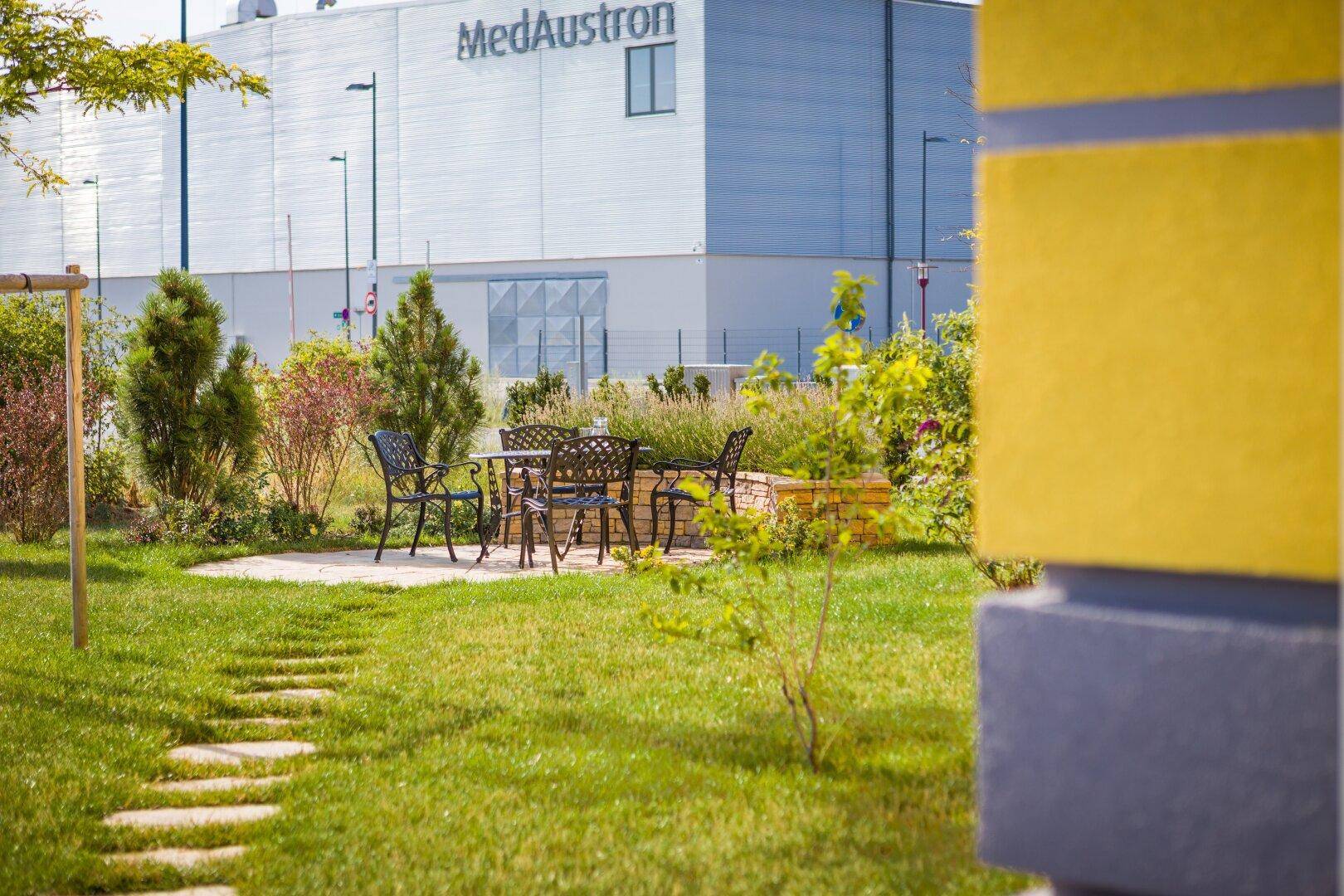 Gartenbereich mit einem runden Sitzplatz und Blick auf ein Industriegebäude im Hintergrund.