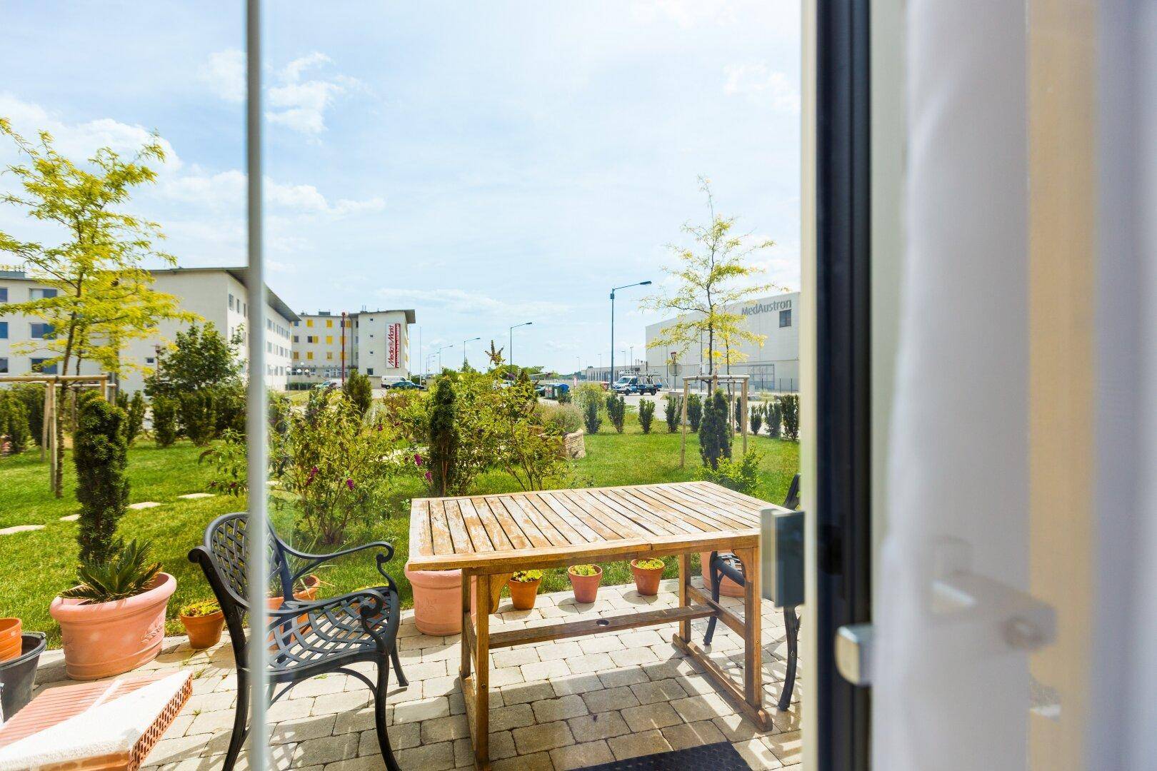 Gepflasterte Terrasse mit Holztisch und Stühlen, umgeben von Grünflächen und Gebäuden.