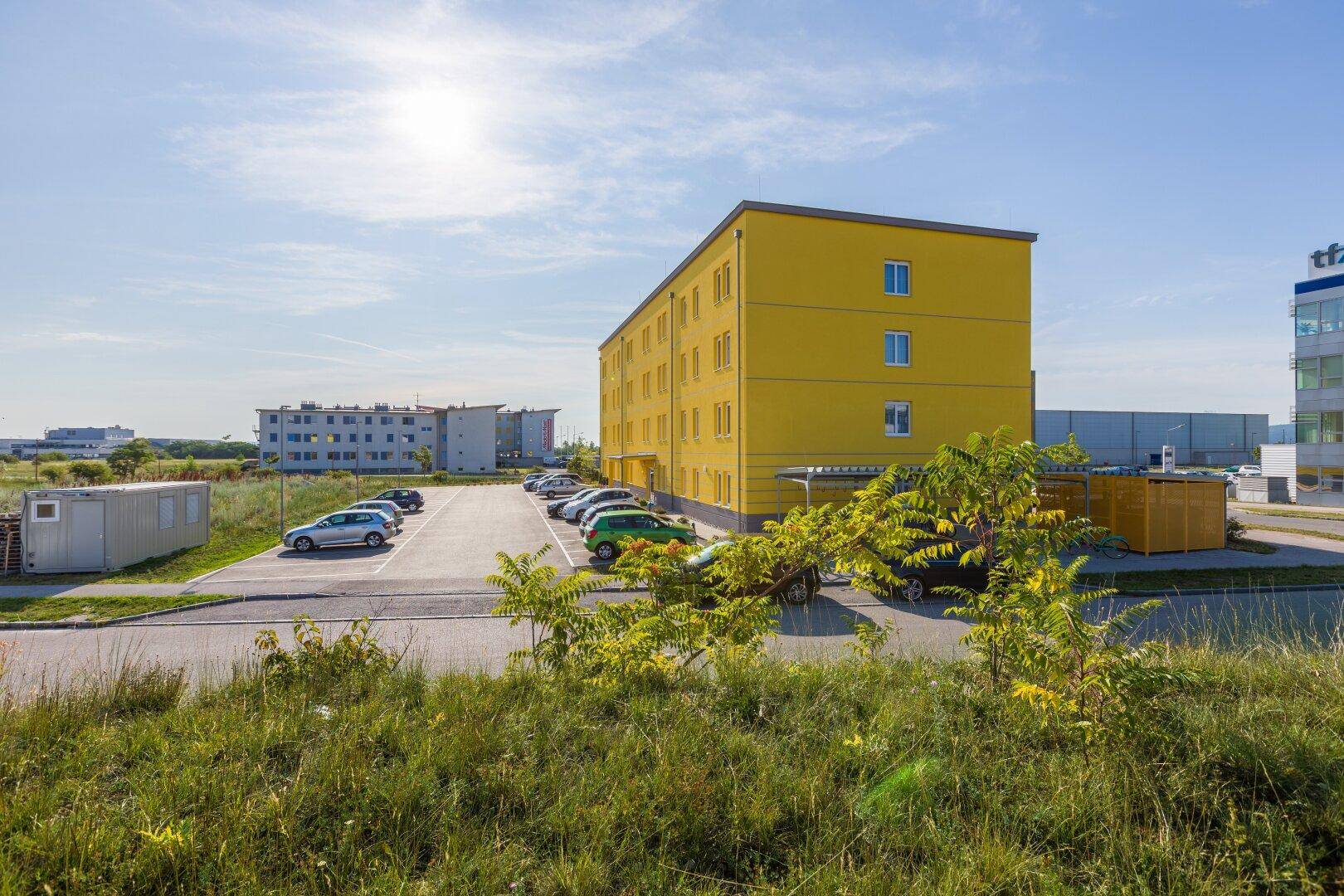 Gelbes Apartmentgebäude mit Parkplatz und Grünflächen unter strahlendem Sonnenschein.