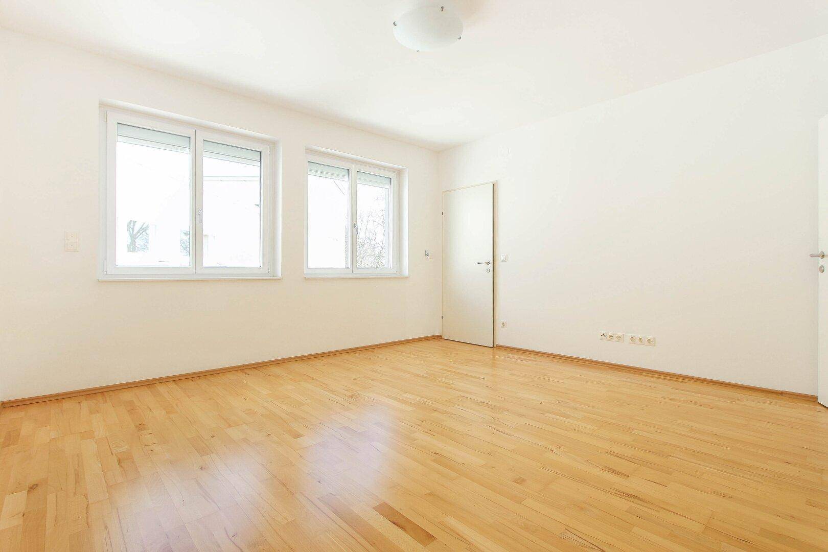 Helles, leeres Zimmer mit Parkettboden und zwei großen Fenstern, ideal als Schlafzimmer.