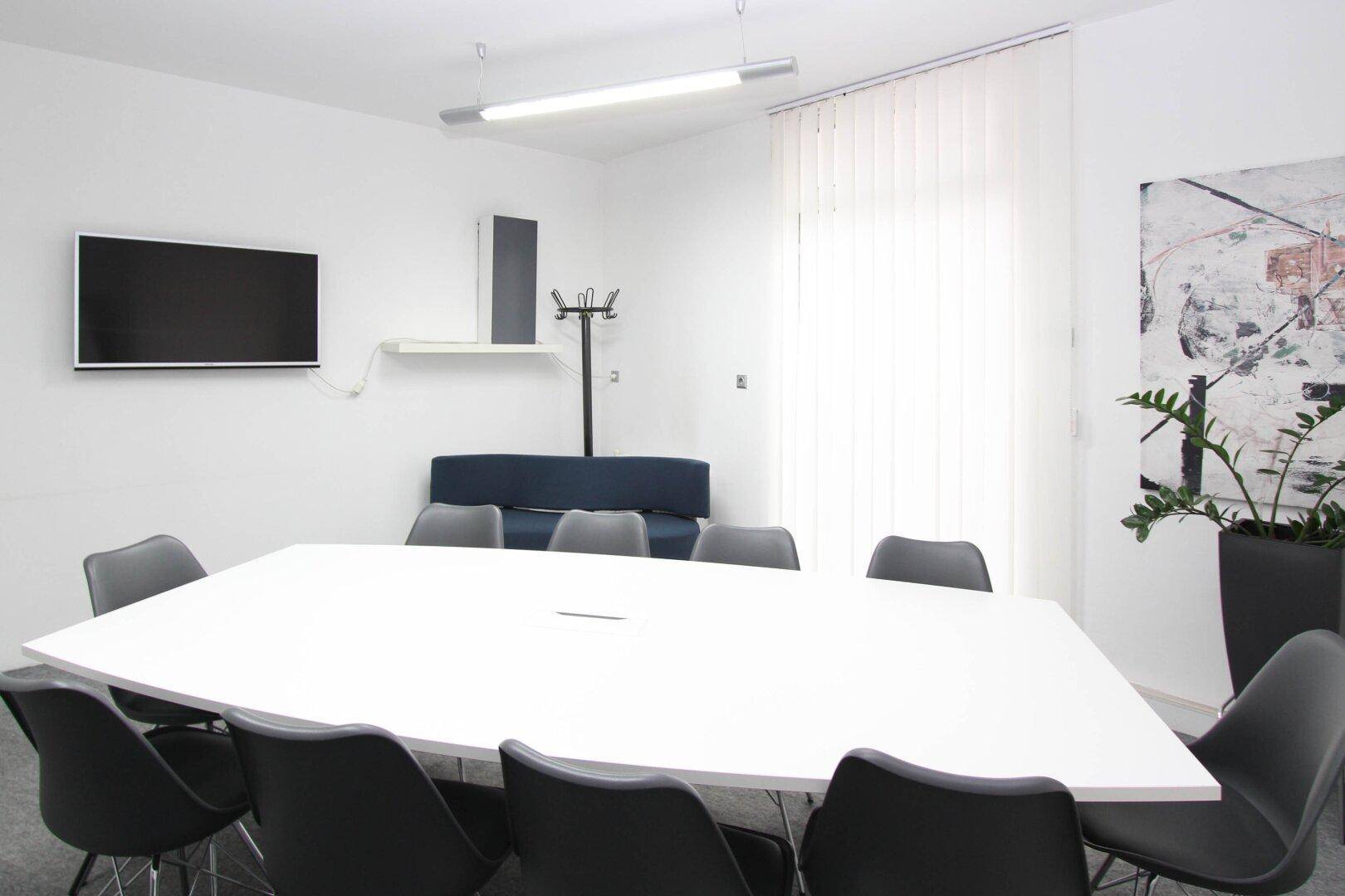 Moderner Konferenzraum mit großem weißen Tisch, bequemen Stühlen und einem Wandfernseher.