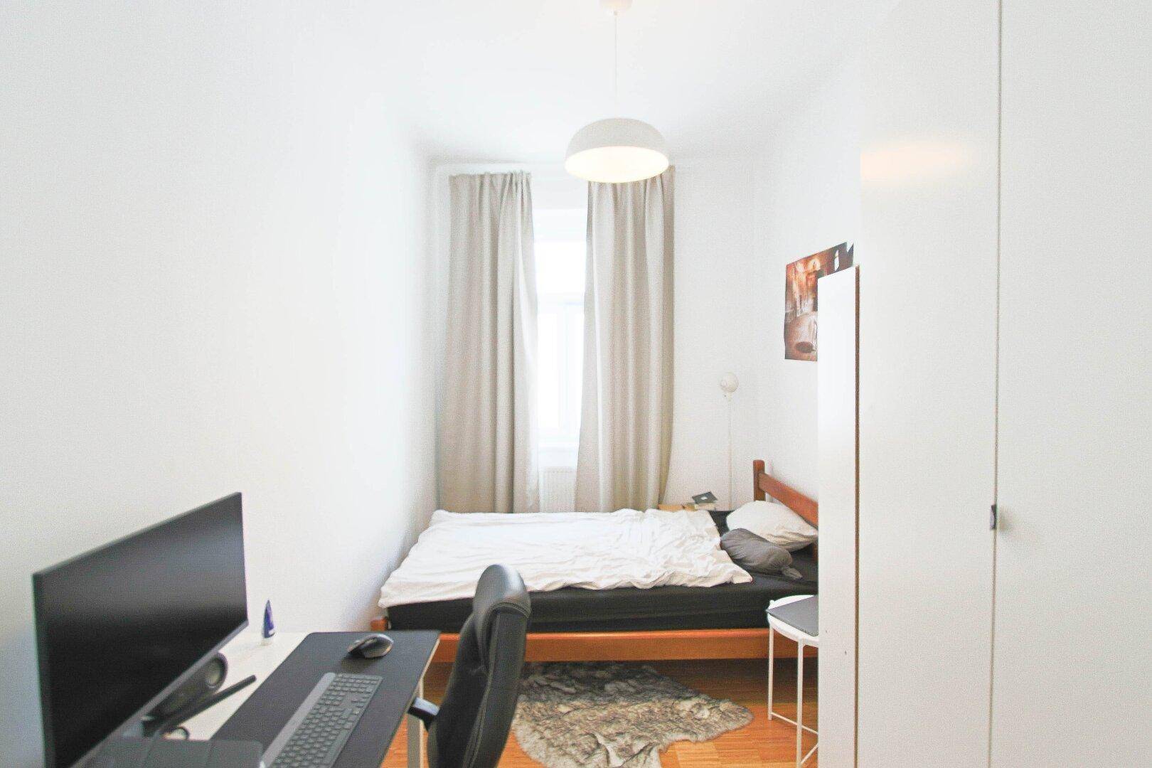 Helles Schlafzimmer mit Bett, Schreibtisch und Fenster, ausgestattet mit einem Teppich.