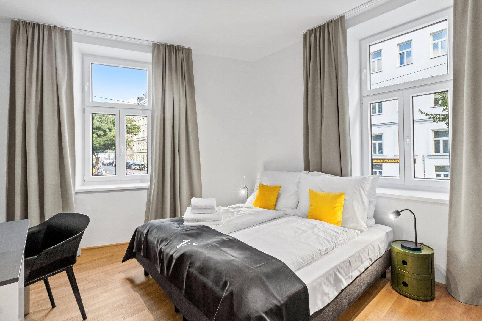 Helles Schlafzimmer mit Doppelbett, Schreibtisch und zwei großen Fenstern mit Stadtblick.