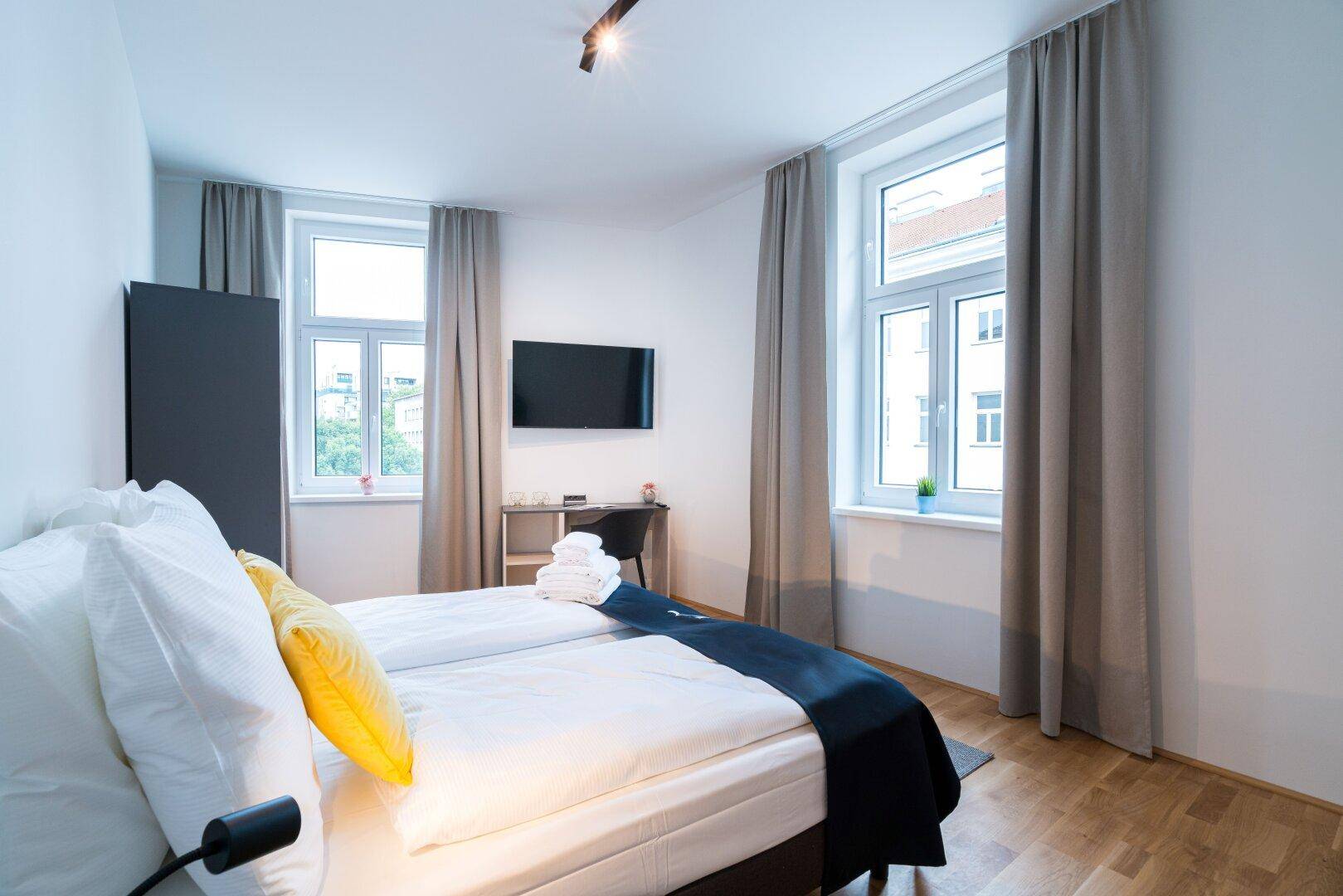 Schlafzimmer mit Doppelbett, Wandfernseher und zwei Fenstern, die viel Licht hereinlassen.