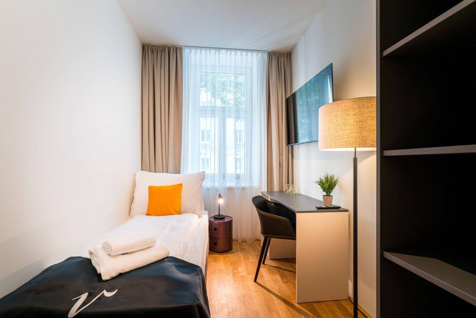 Kleines, helles Schlafzimmer mit Einzelbett, Schreibtisch und Stehlampe, funktional eingerichtet.