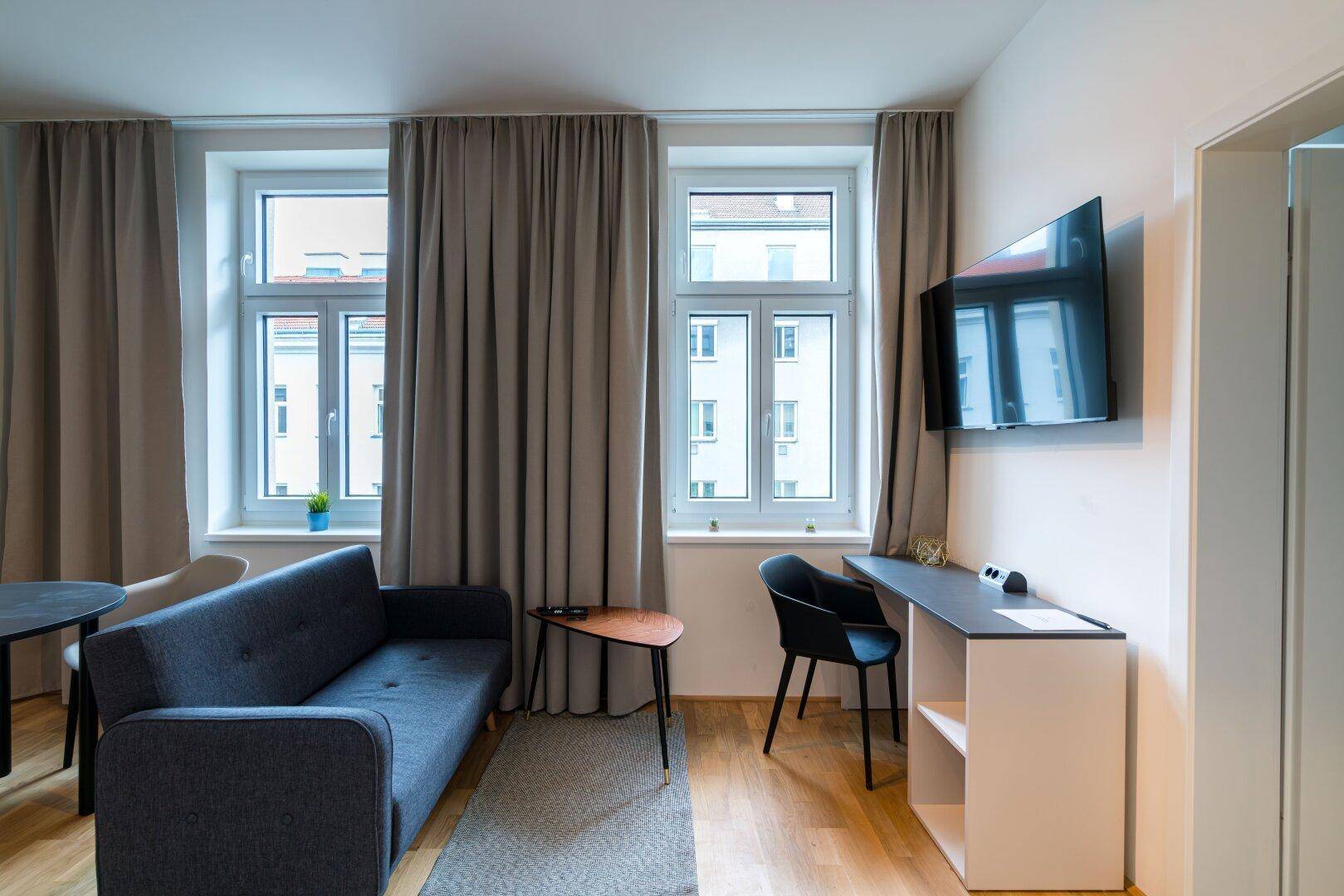 Heller Wohnbereich mit Sofa, Schreibtisch und großen Fenstern mit Vorhängen.
