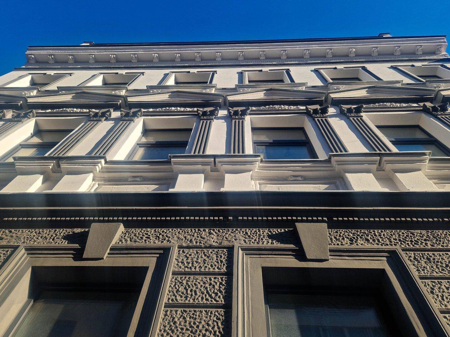 Detailansicht der historischen Fassade mit Stuckelementen und großen Fenstern unter strahlend blauem Himmel.