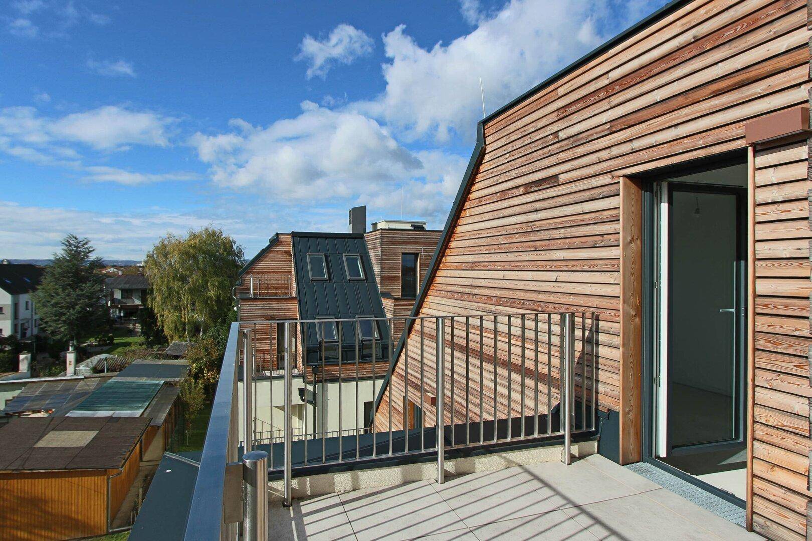 Balkon mit Holzfassade und Blick auf die umliegende Wohngegend unter blauem Himmel.
