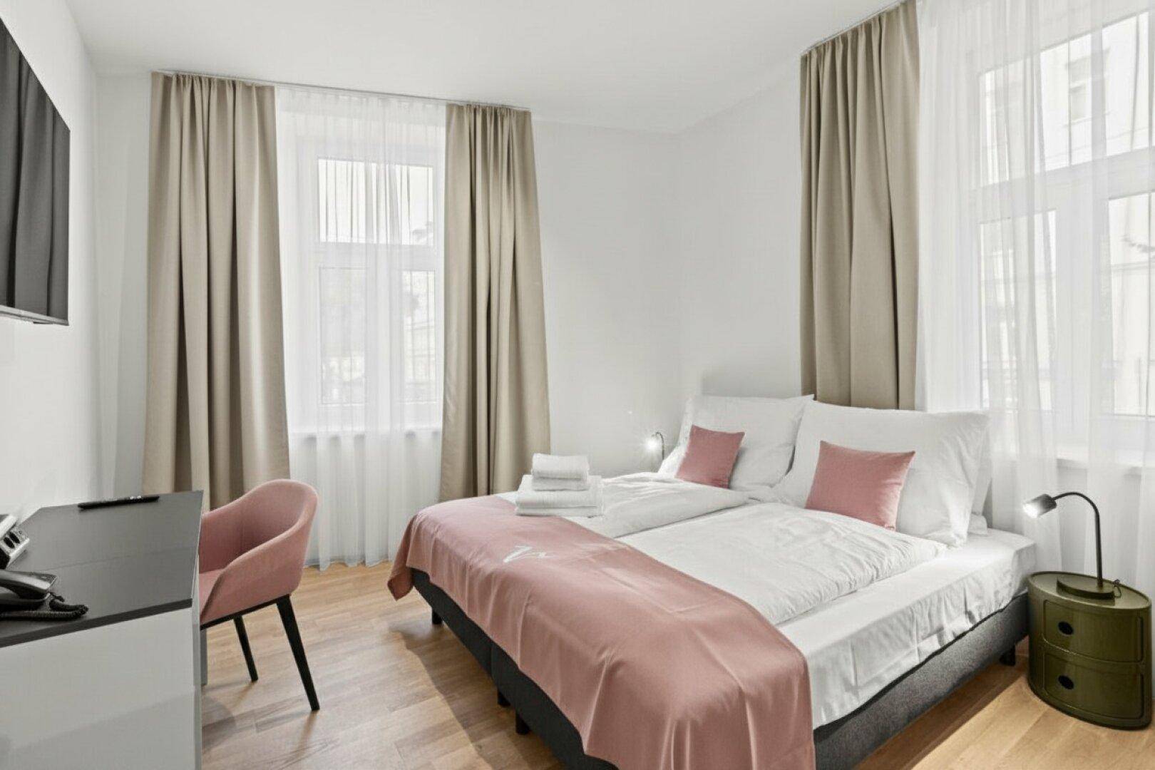 Helles Schlafzimmer mit Doppelbett, Schreibtisch und zwei Fenstern, modern eingerichtet.