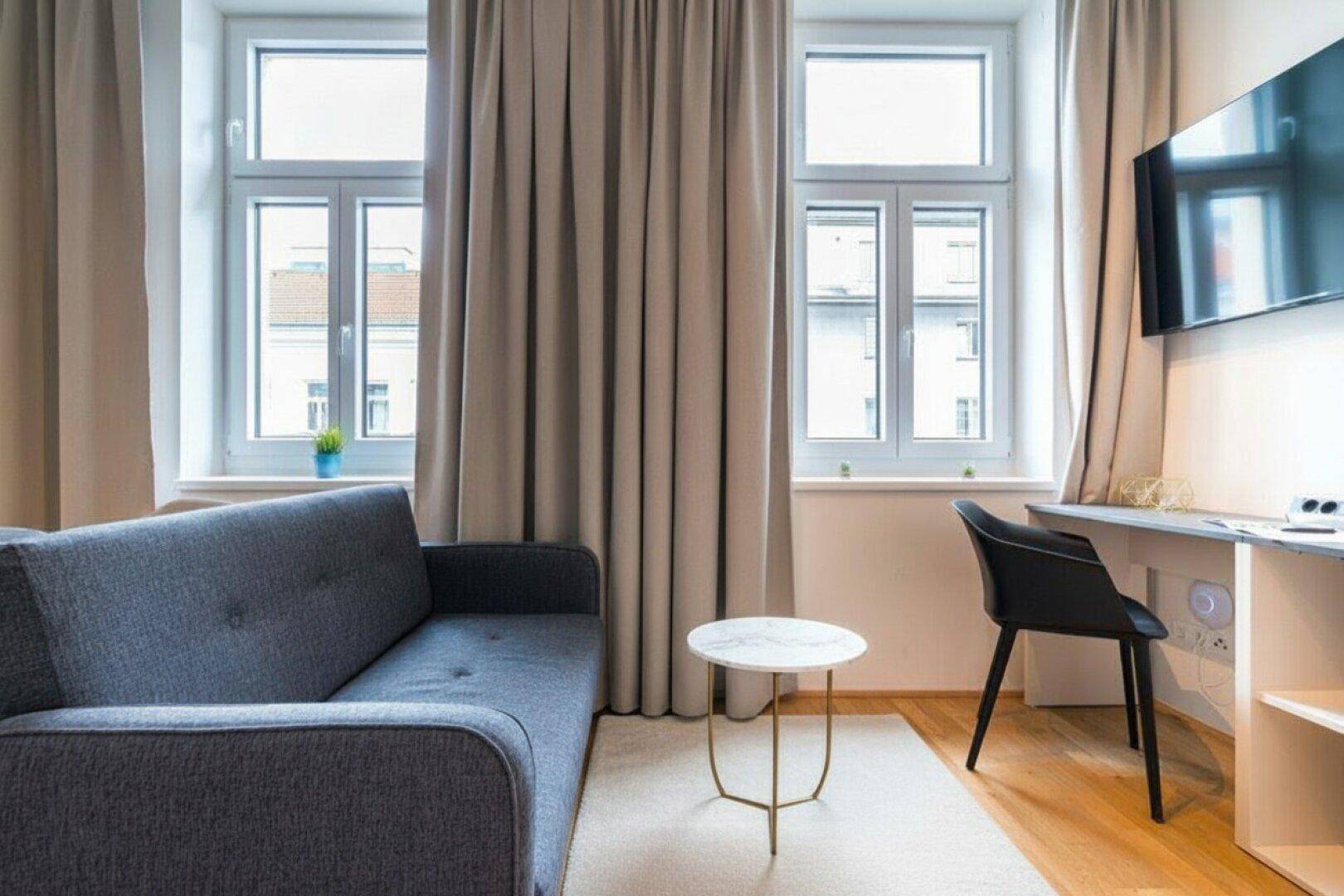Gemütliches Wohnzimmer mit Sofa, Schreibtisch und großen Fenstern für viel Tageslicht.