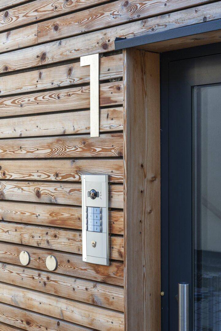 Eingangsbereich mit Holzfassade, moderner Gegensprechanlage und Hausnummer.