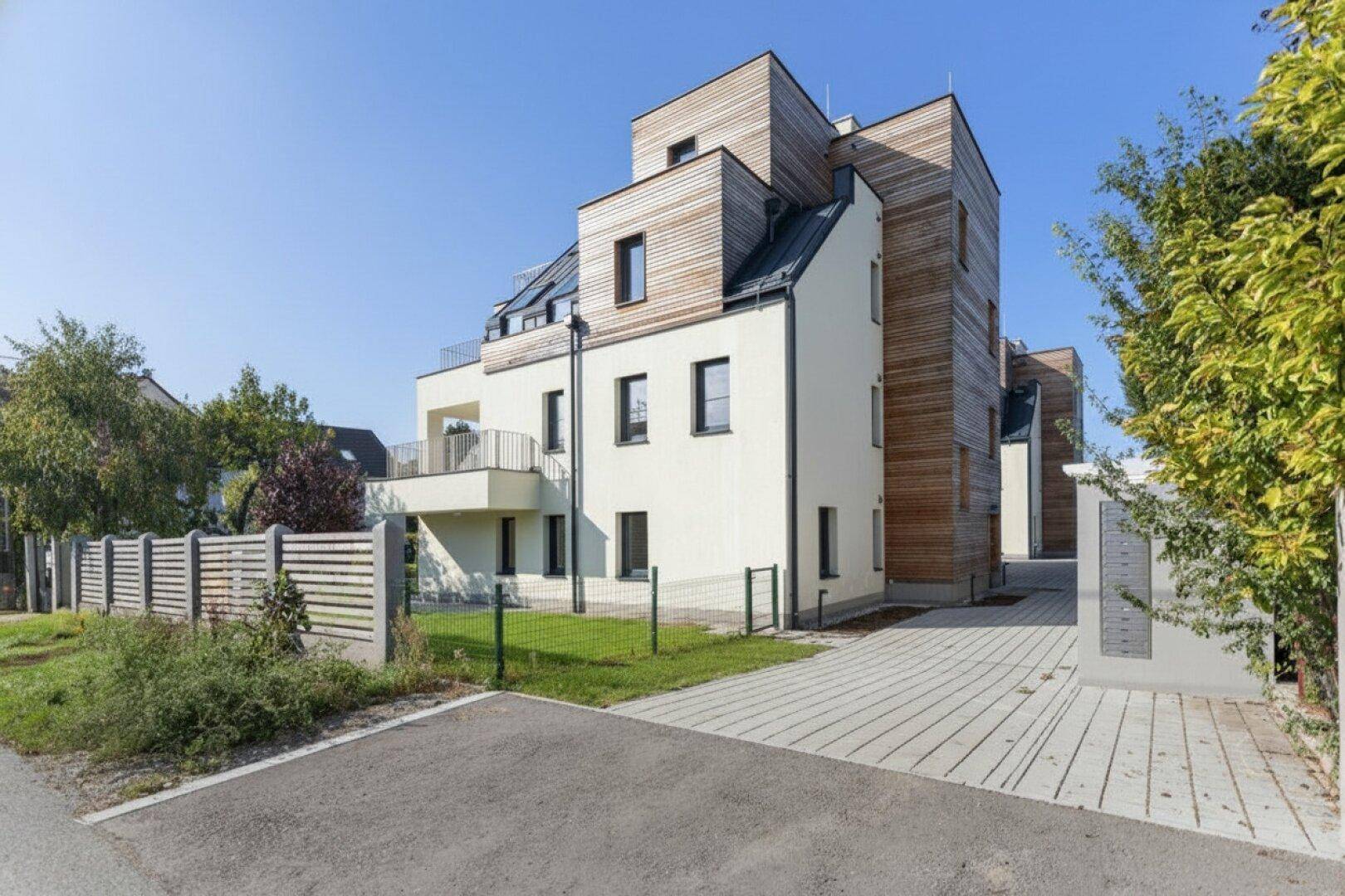 Modernes Mehrfamilienhaus mit Holzfassade und hellen Putzflächen, umgeben von Grün.