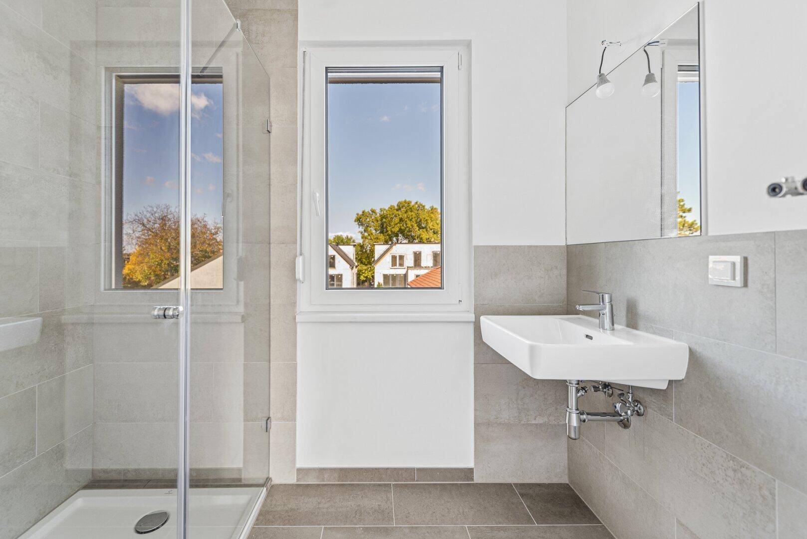 Modernes Badezimmer mit Dusche, Waschbecken und Fenster, hell gefliest in Grautönen.