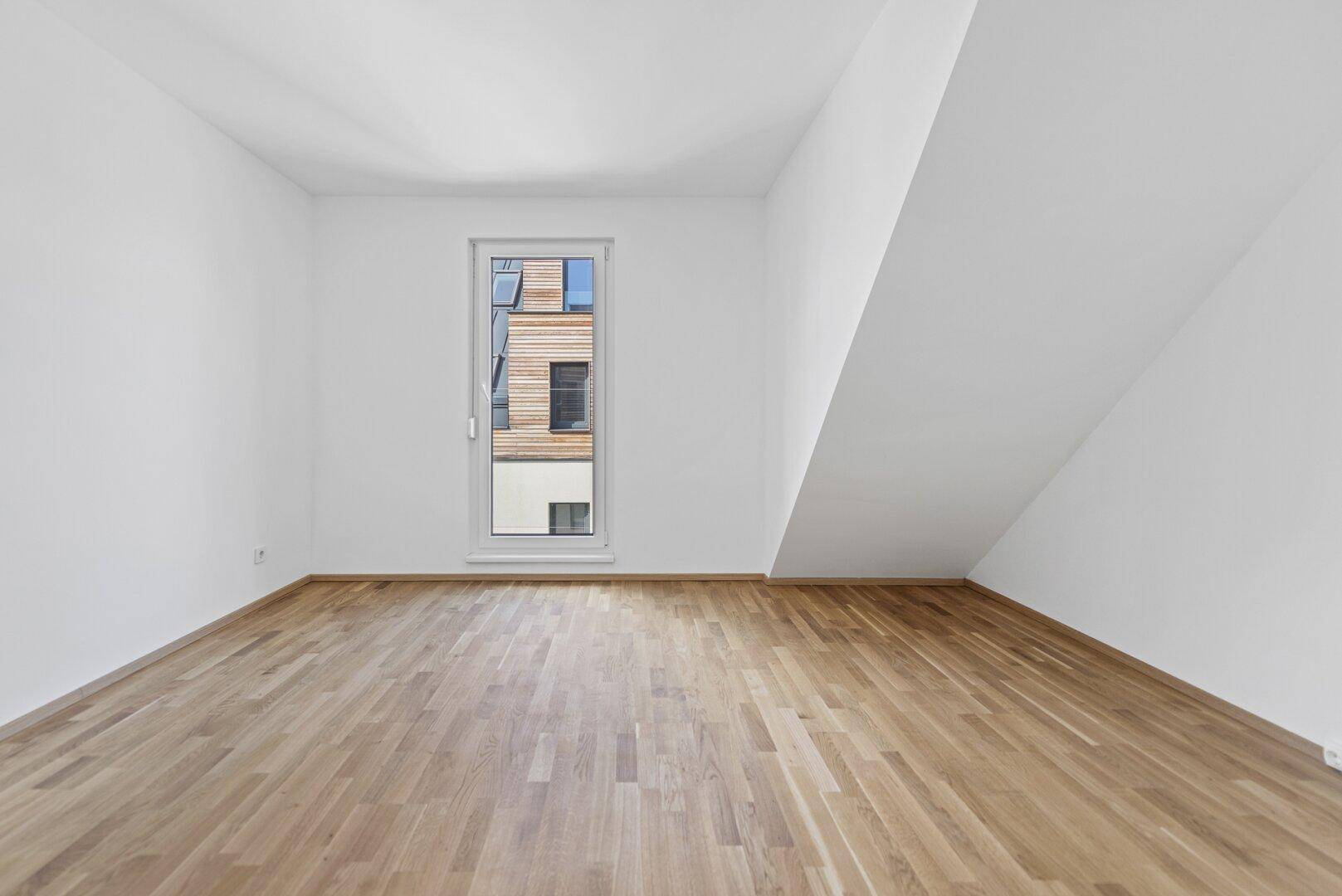 Schlichtes Zimmer mit Holzboden und einem hohen Fenster, das den Blick nach draußen ermöglicht.