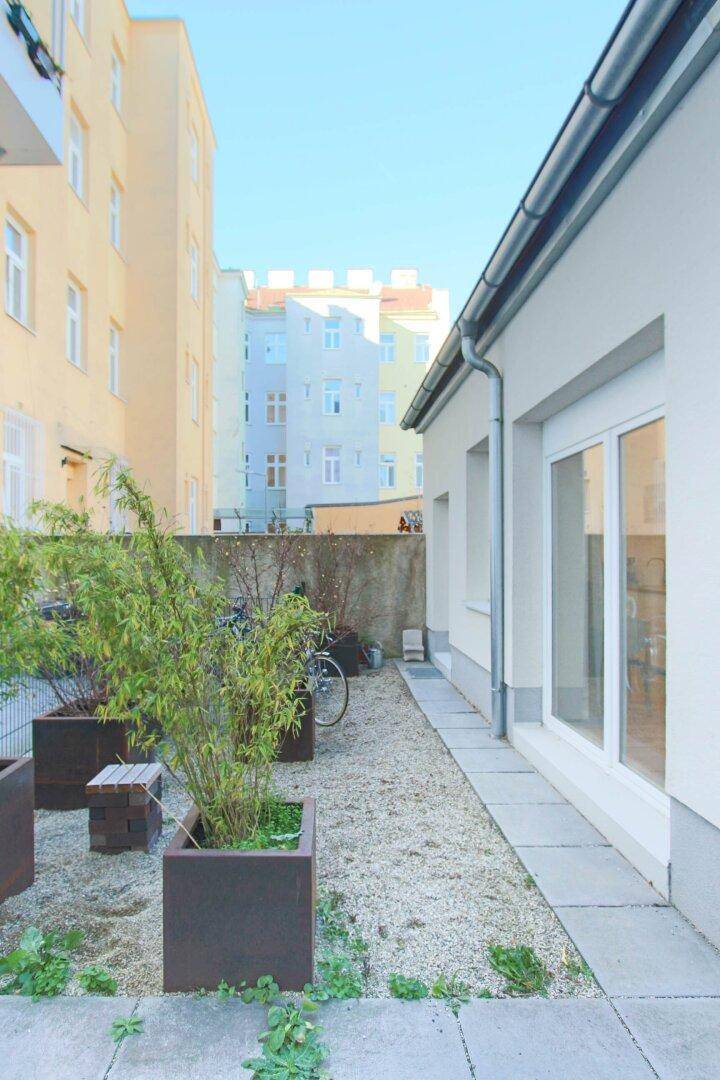 Ruhiger Gartenbereich mit Kies und Pflanzenkübeln, umgeben von städtischen Gebäuden und einer Mauer.