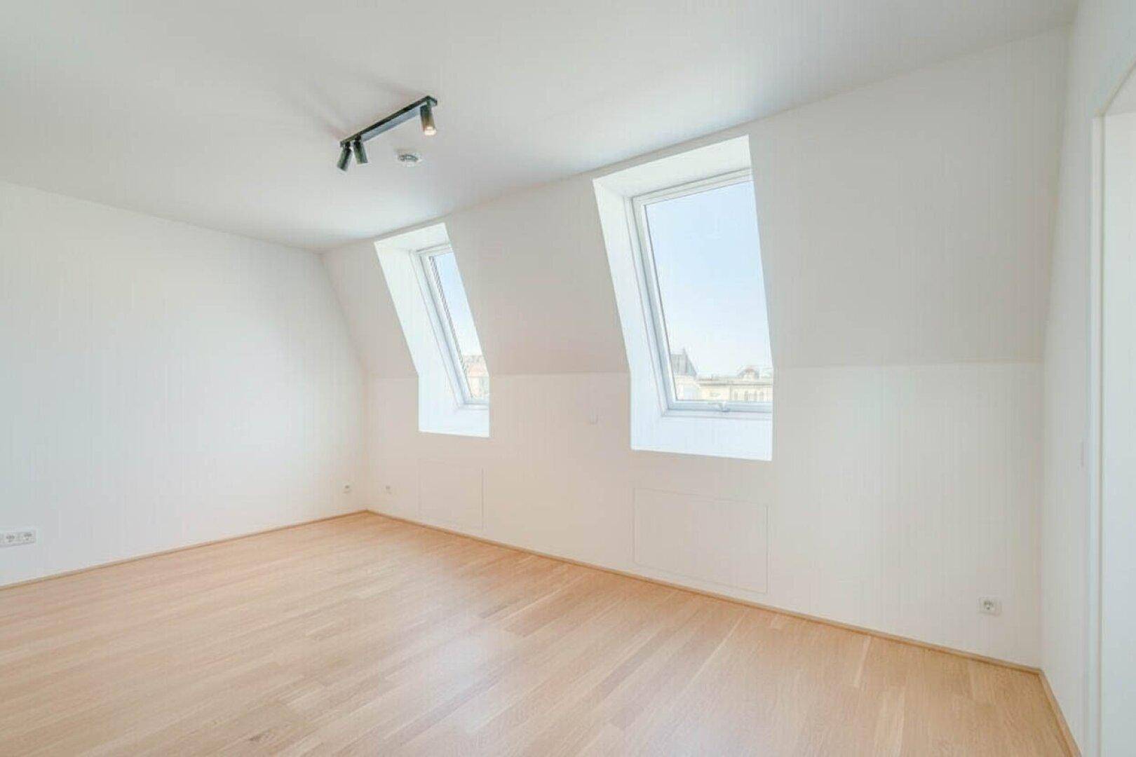 Heller Raum im Dachgeschoss mit zwei Dachfenstern, Holzfußboden und schrägen Wänden.