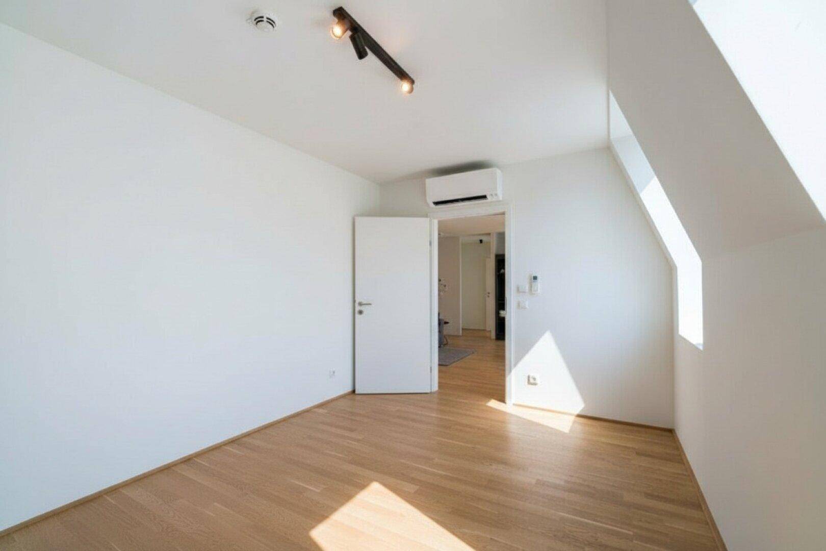 Helles Zimmer mit Holzfußboden, Dachschräge, Klimaanlage und Zugang zu einem weiteren Raum.