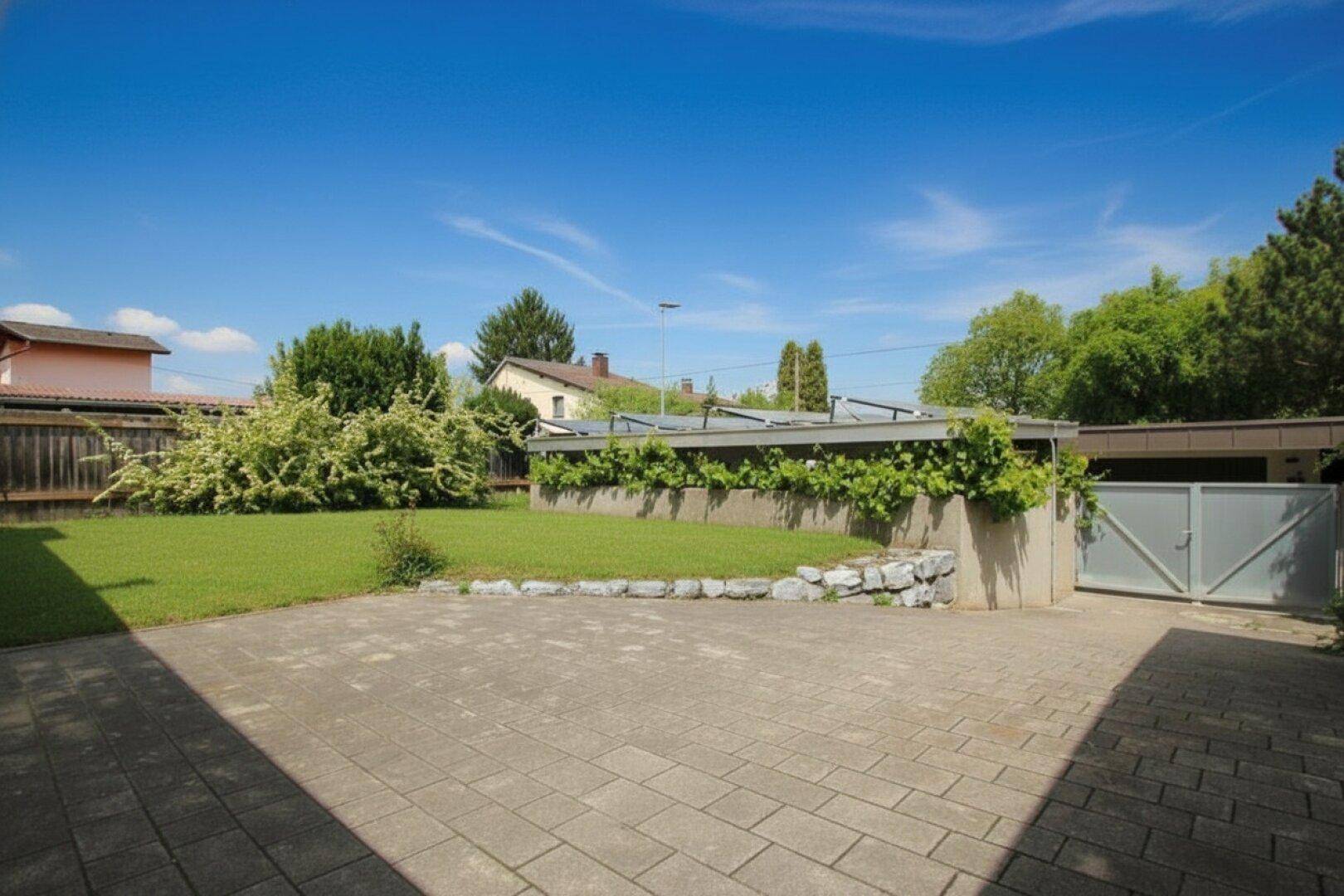 Gepflasterter Vorplatz mit niedriger Steinmauer, begrünten Rankpflanzen und Rasenfläche.