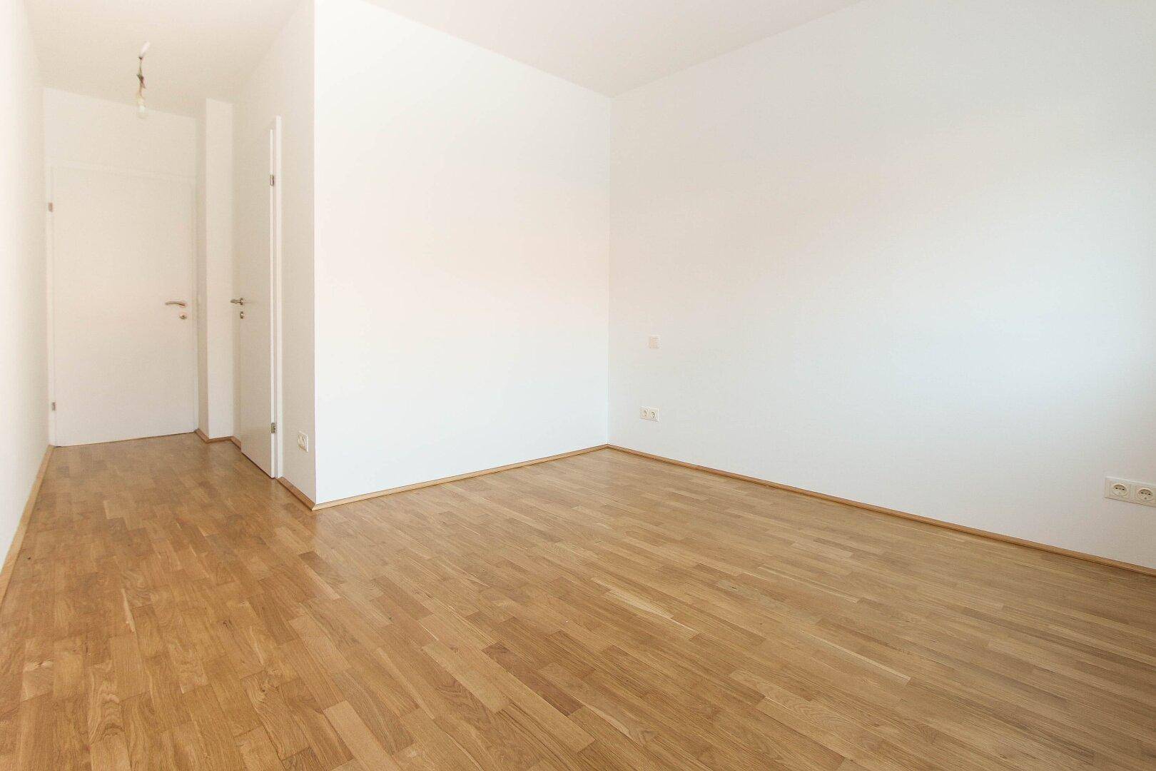 Leeres Zimmer mit Holzboden, weißen Wänden und einer Tür zum Flur.
