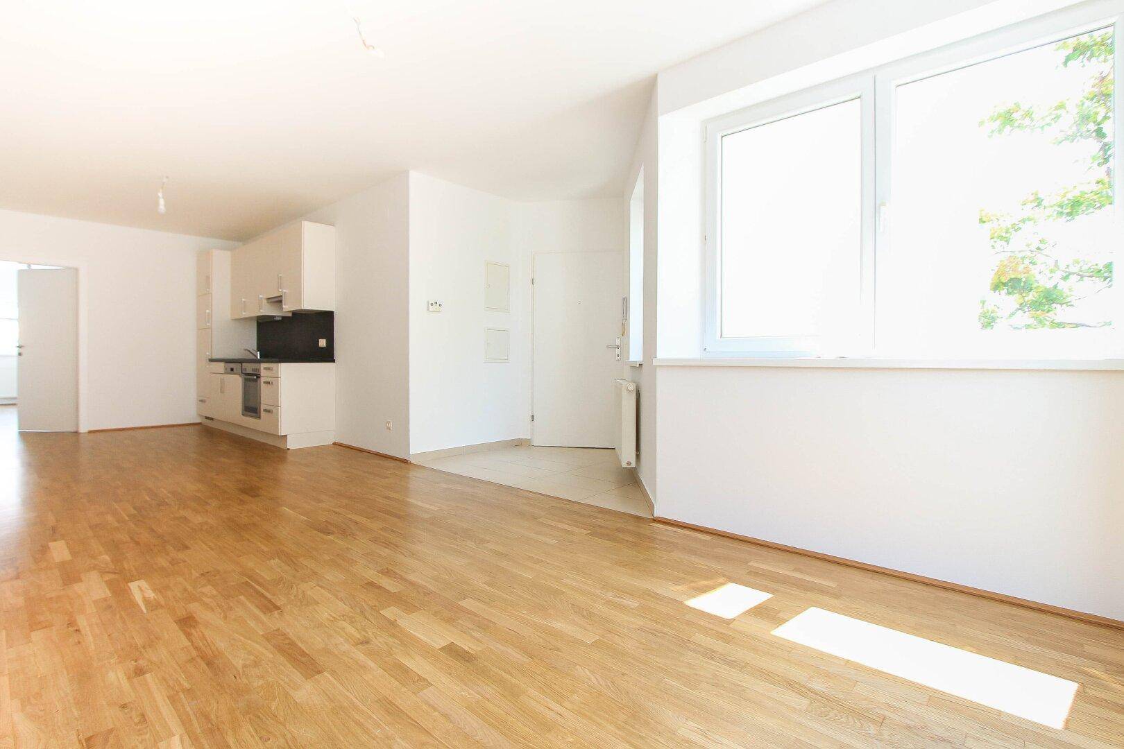 Heller Wohnbereich mit Holzboden, großen Fenstern und einer offenen Küchenzeile im Hintergrund.