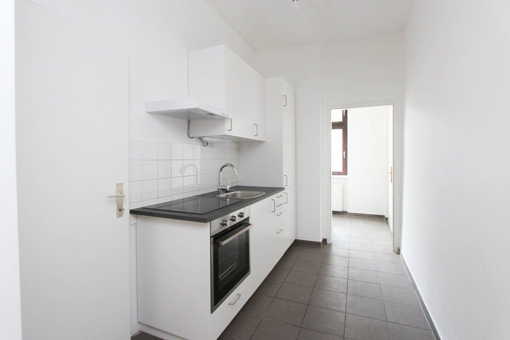 Funktionale Küche mit weißen Einbauschränken, Herd und Spüle, mit Zugang zu einem weiteren Zimmer.