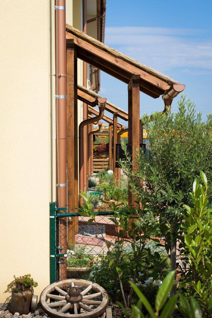 Seitenansicht eines Hauses mit hölzerner Pergola-Struktur und üppiger Bepflanzung im Vorgartenbereich.