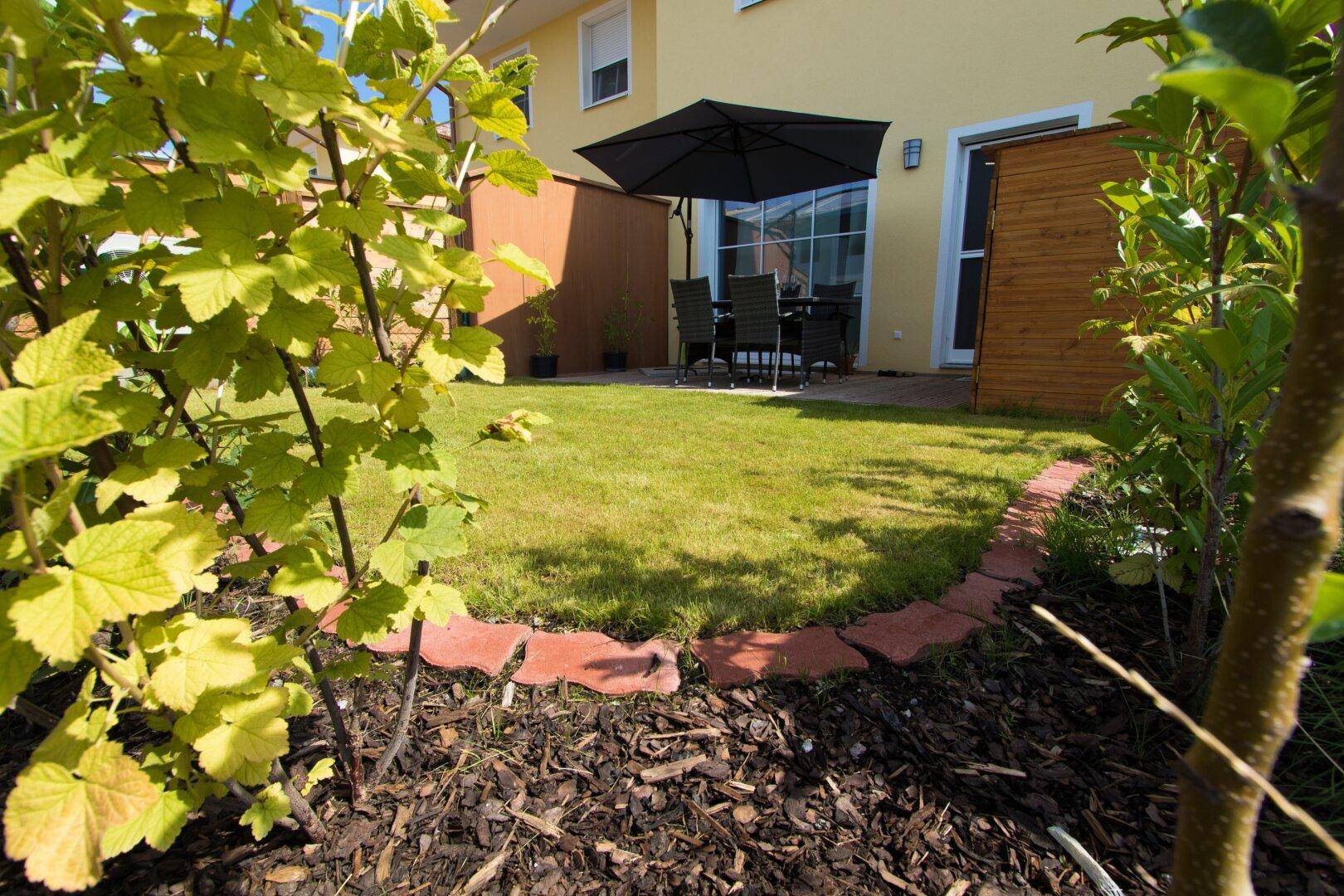 Kleiner, gepflegter Garten mit Rasenfläche, einer Terrasse und Gartenmöbeln unter einem Sonnenschirm.