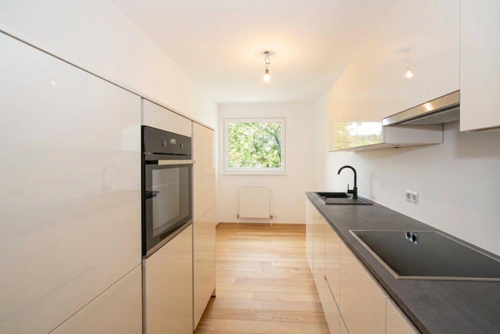Helle Küche mit Fenster, weißen Hochglanzfronten, dunkler Arbeitsplatte und Induktionskochfeld.