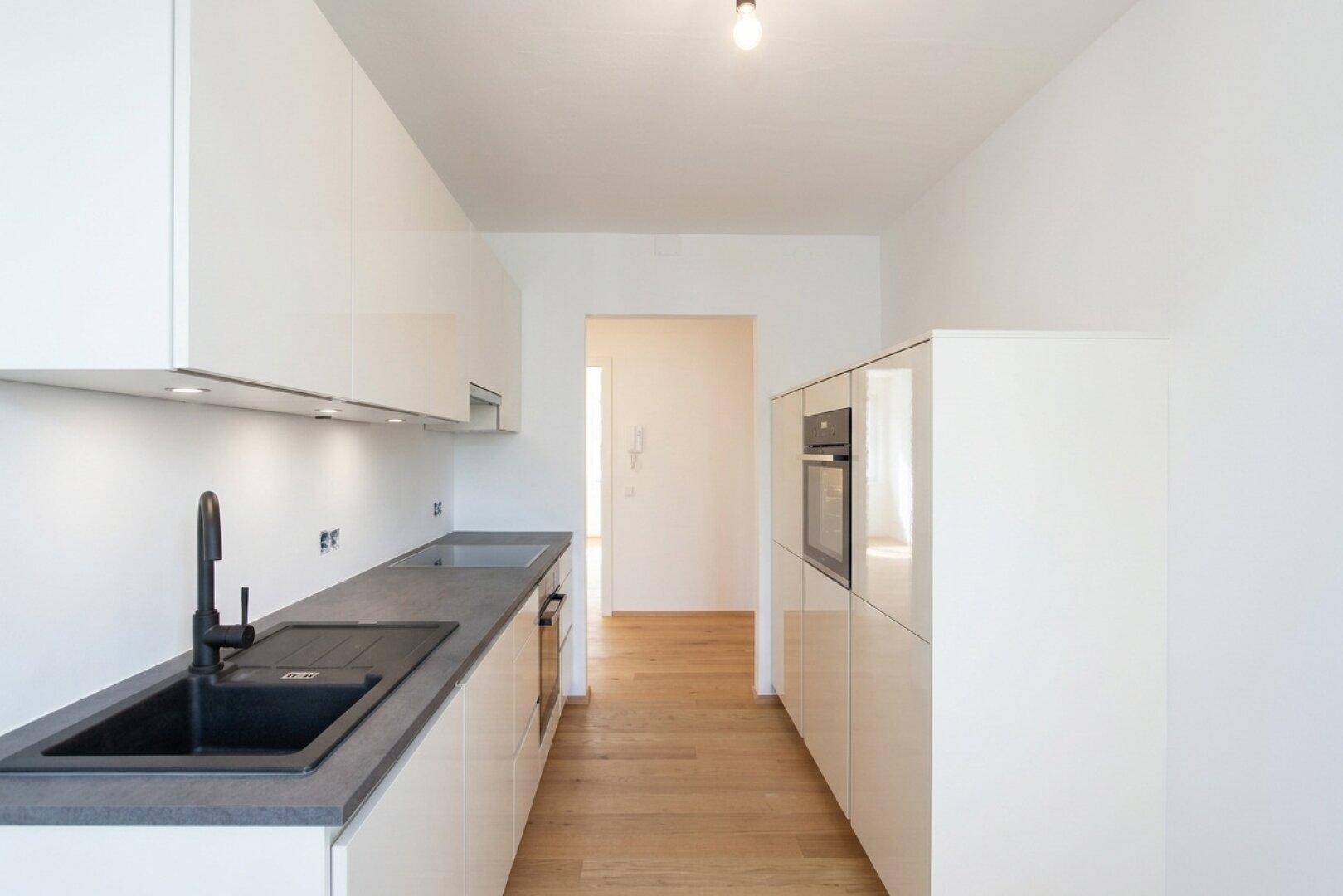 Moderne, schmale Küche mit weißen Hochglanzschränken, dunkler Arbeitsplatte und integrierten Geräten.