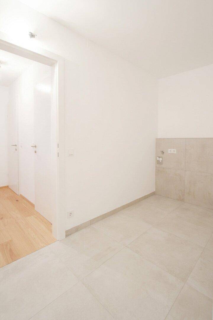 Leerer Raum mit hellgrauen Fliesen und Anschlüssen an der Wand, möglicherweise für eine Waschmaschine.