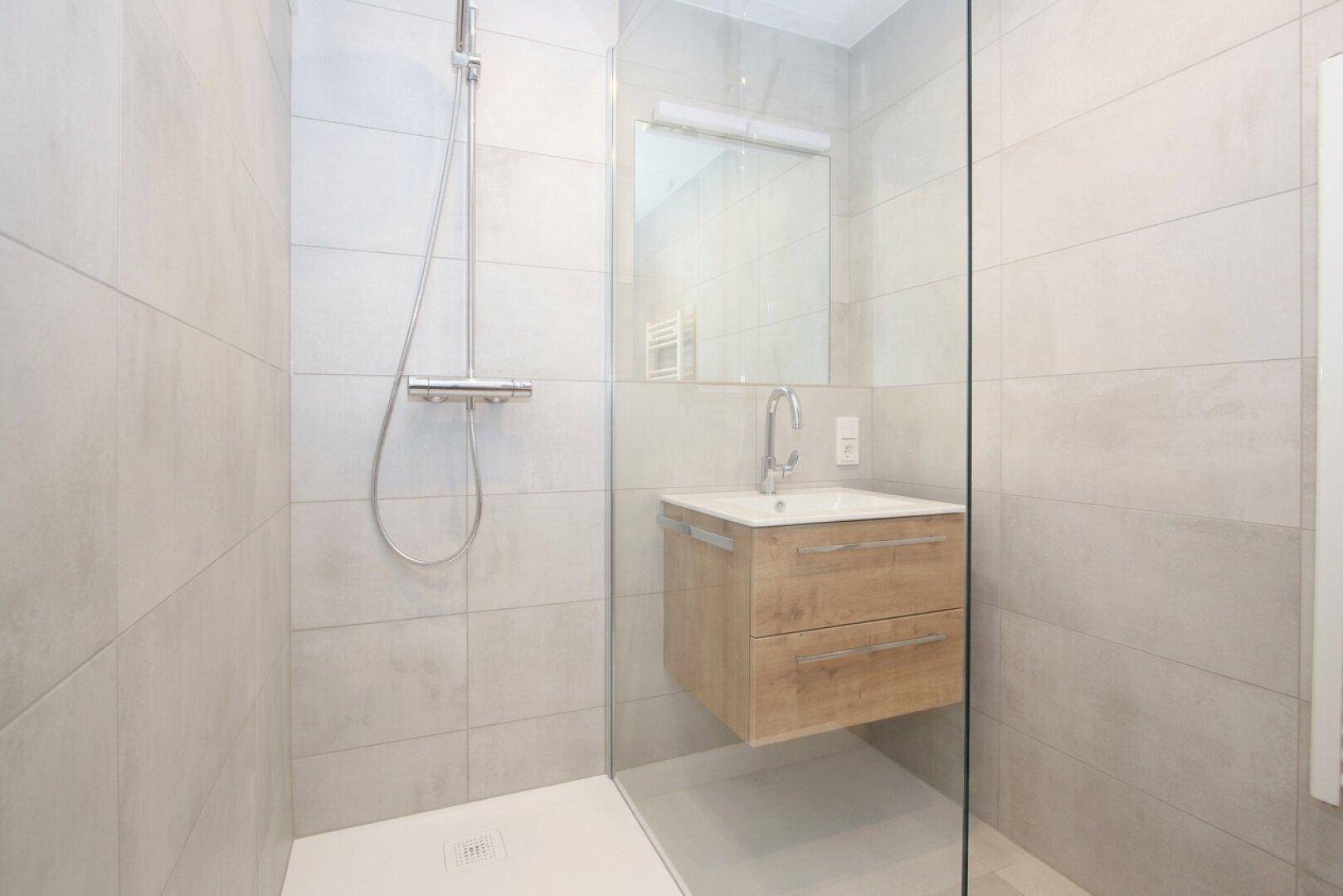 Modernes Badezimmer mit begehbarer Dusche, Holzwaschtisch und hellgrauen Fliesen an Wänden und Boden.