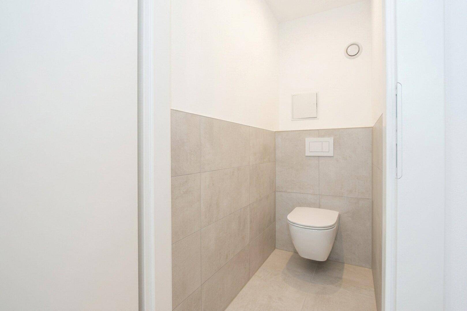 Kleines, separates WC mit wandhängender Toilette und halbhohen, hellgrauen Wandfliesen.